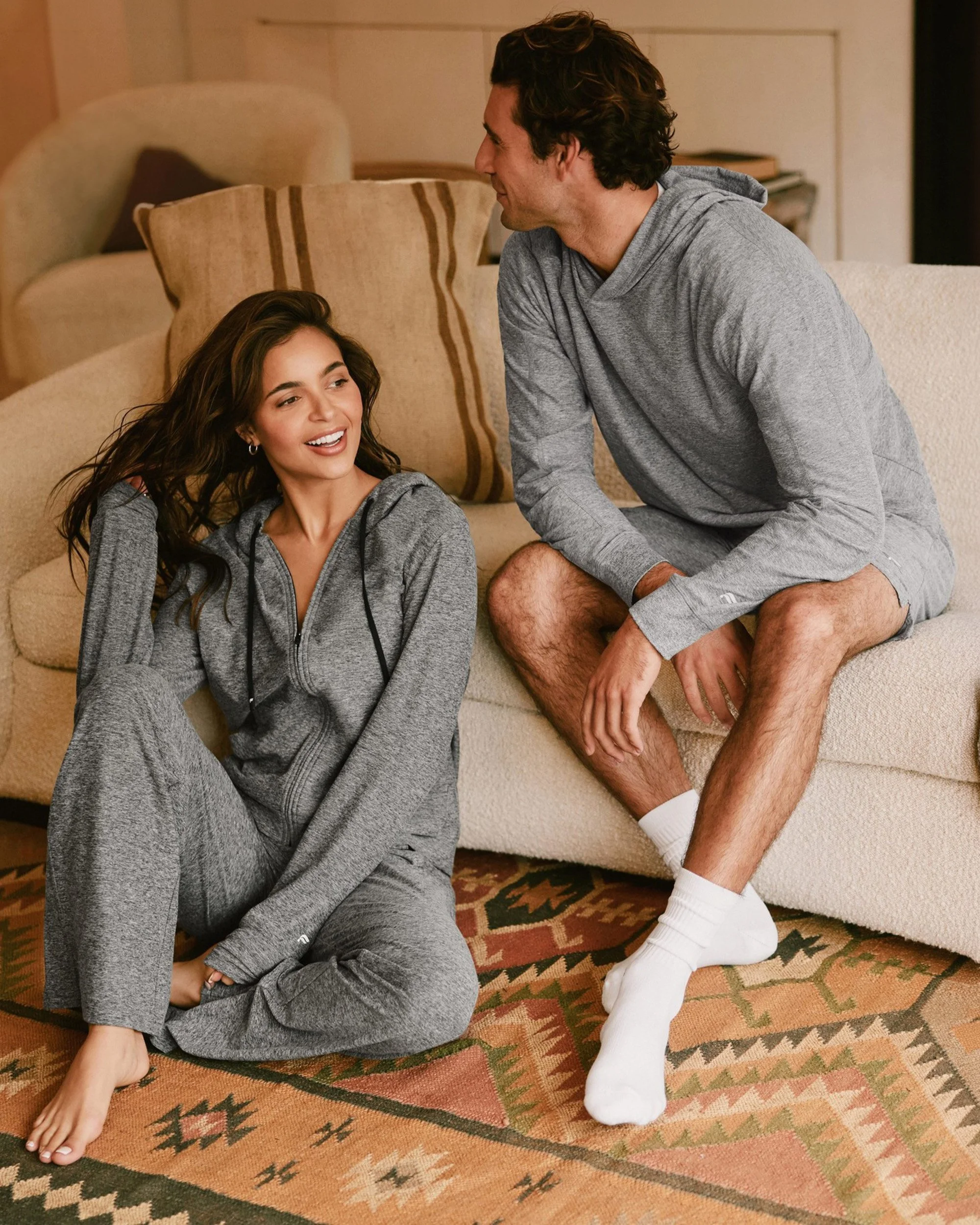 Art director on-figure e-commerce shoot, Los Angeles. A young woman sitting on the floor, smiling, talking to a young man sitting on a sofa. Both are in matching gray loungewear.