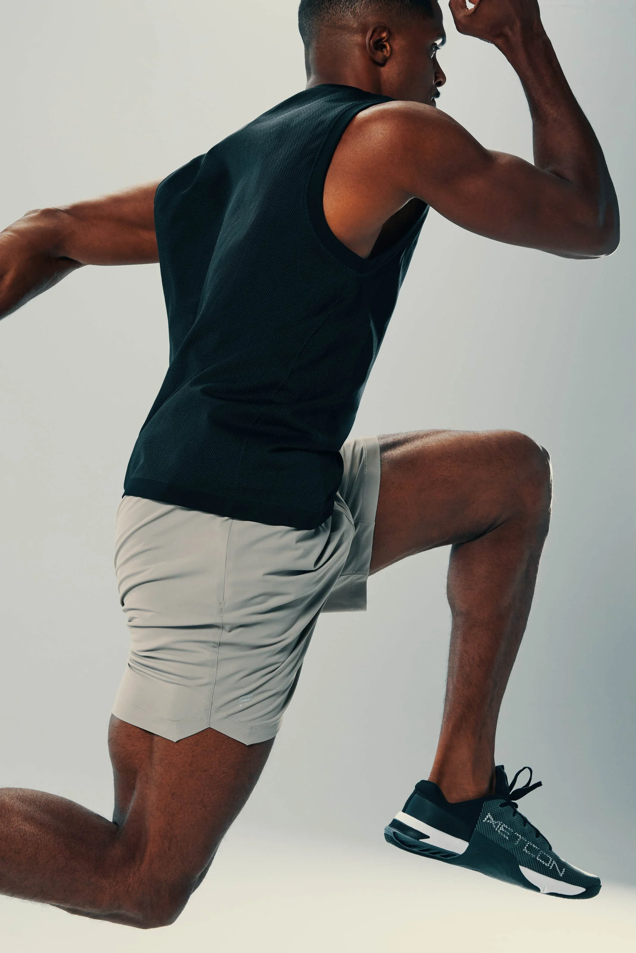 A man in athletic gear is in a running or sprinting pose, wearing a black sleeveless shirt, beige shorts, and black running shoes.