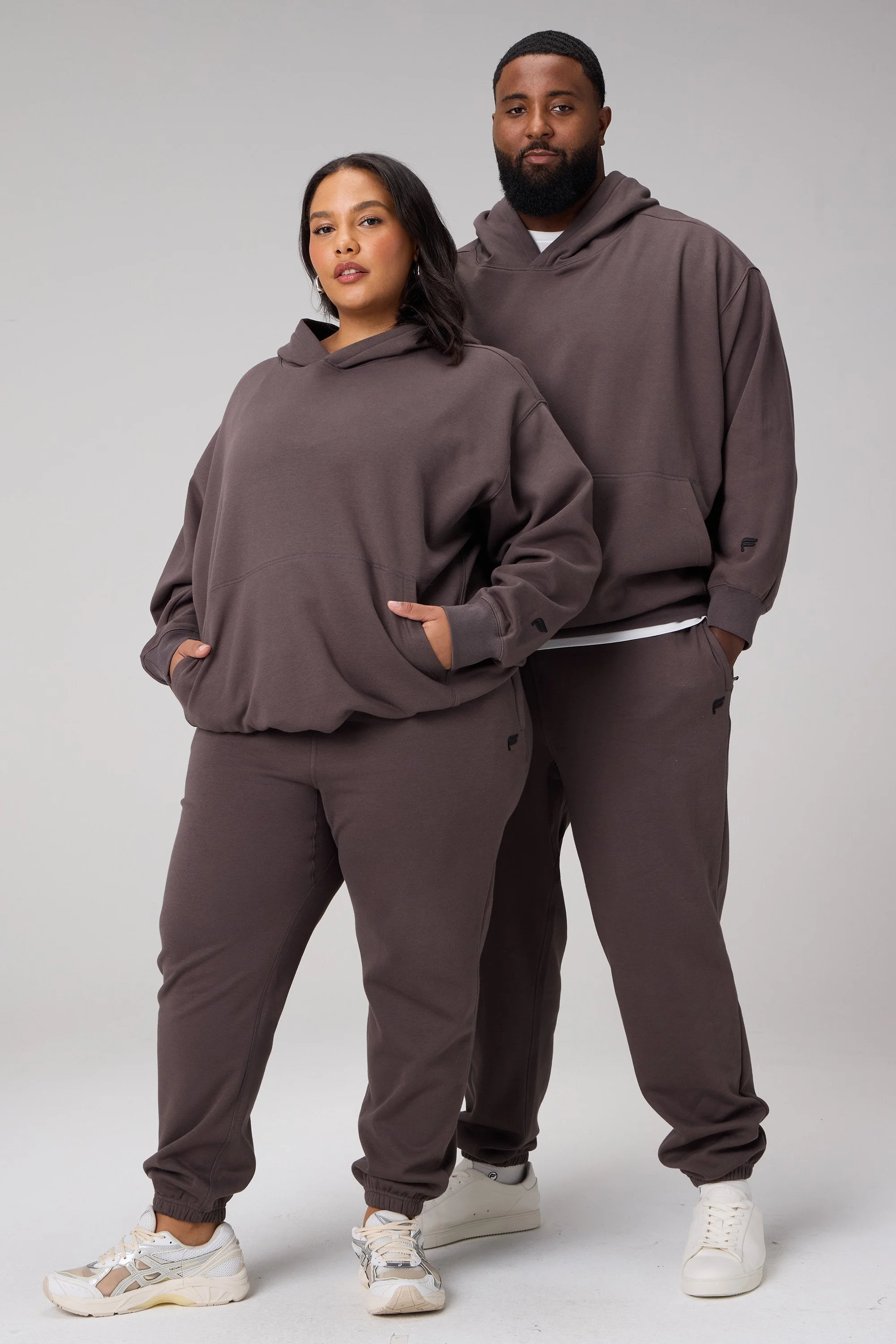 Two individuals, a woman and a man, wearing matching dark gray hoodies and sweatpants, standing against a plain light gray background.