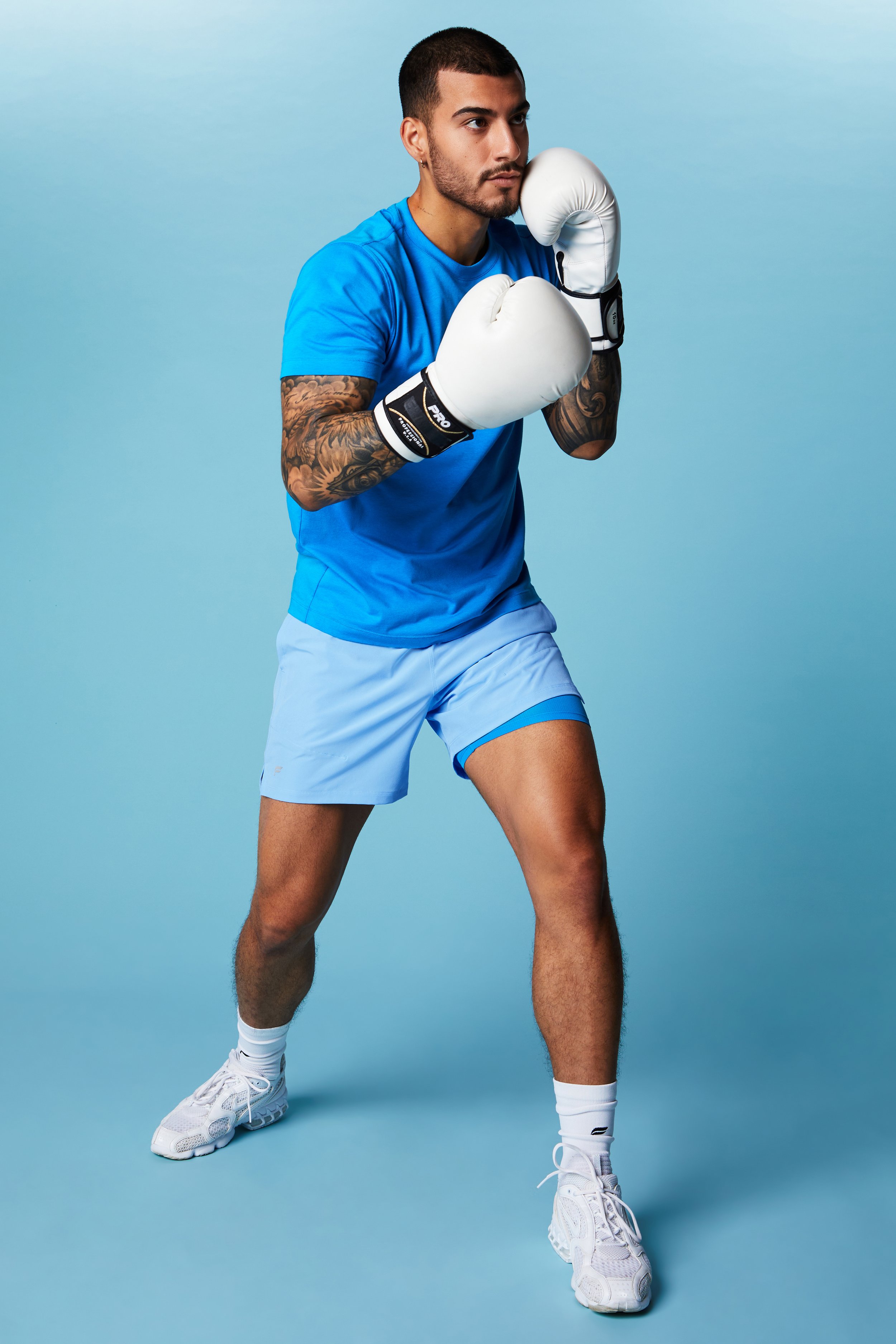 A young man in blue athletic clothing and white boxing gloves posing in a boxing stance against a light blue background.