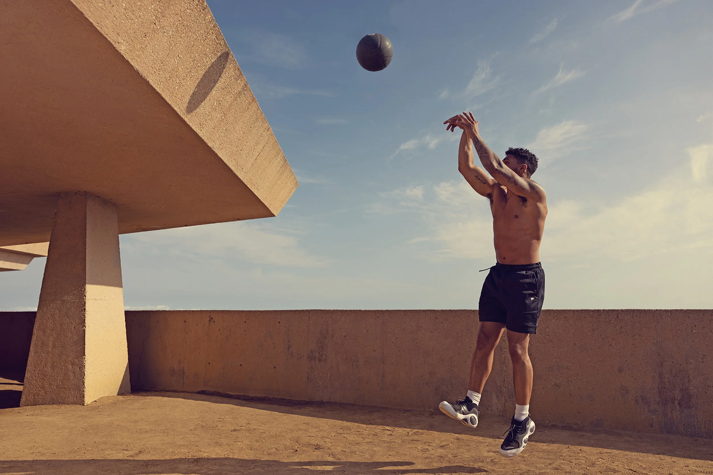 Art director on-figure e-commerce shoot, Los Angeles. A shirtless man wearing black shorts, white socks, and black sneakers is jumping and preparing to hit a black ball with his hands on an outdoor concrete terrace with a blue sky background.