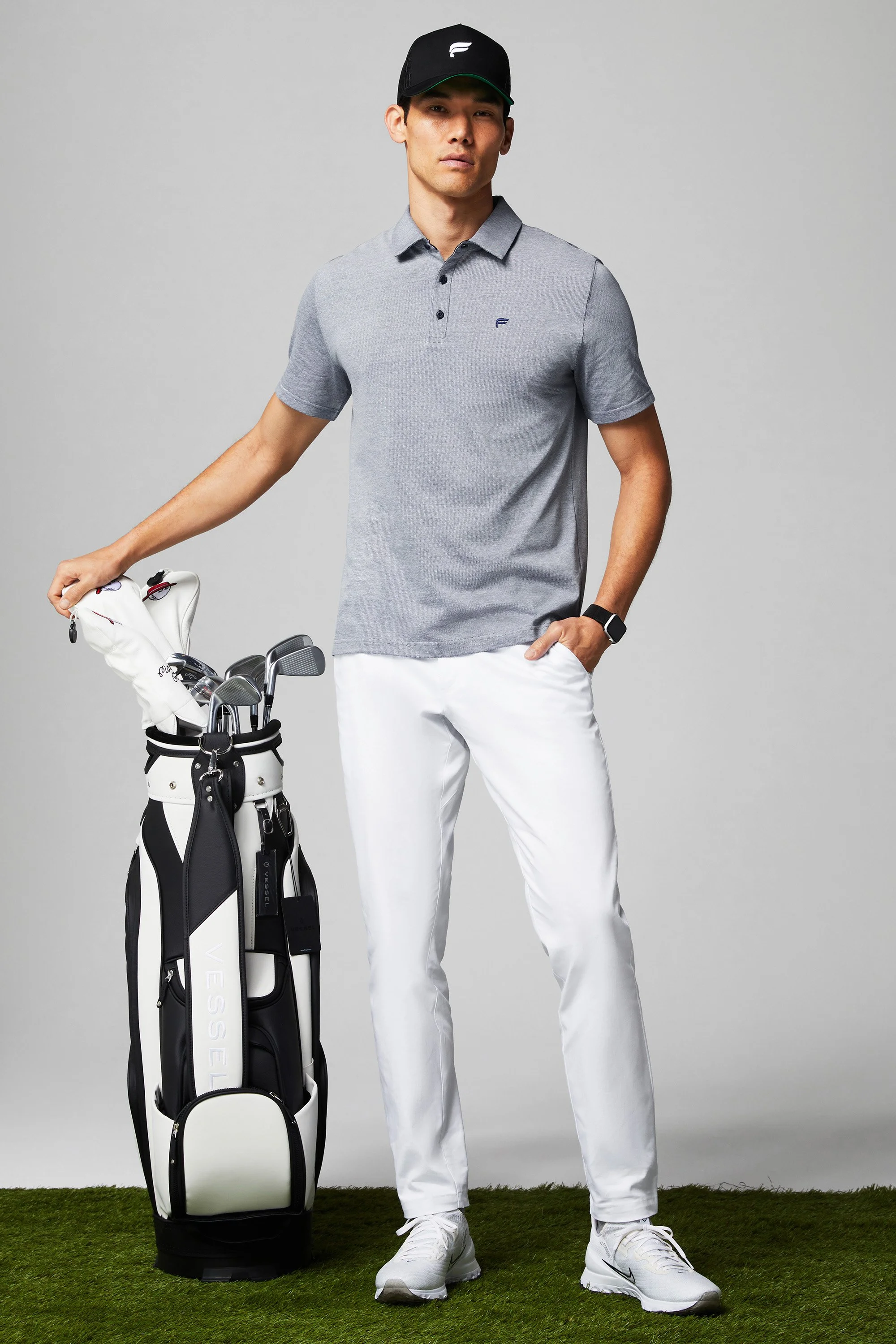 A man dressed in a gray polo shirt and white pants stands next to a golf bag filled with clubs, wearing a black cap and white golf shoes, on a green surface.