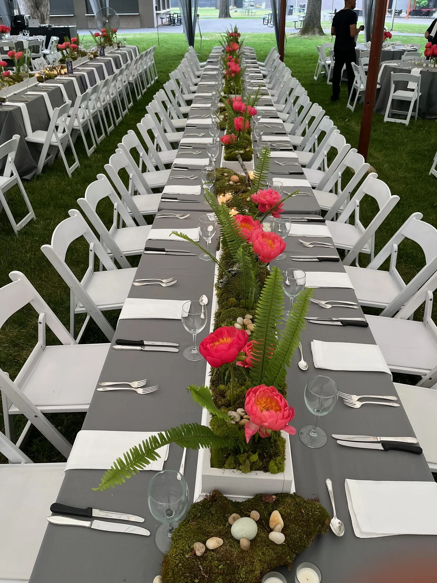 Long dining table decorated with pink flowers, ferns, and moss, set with white napkins, silverware, and empty glasses, in an outdoor tented setting for a gathering or event.