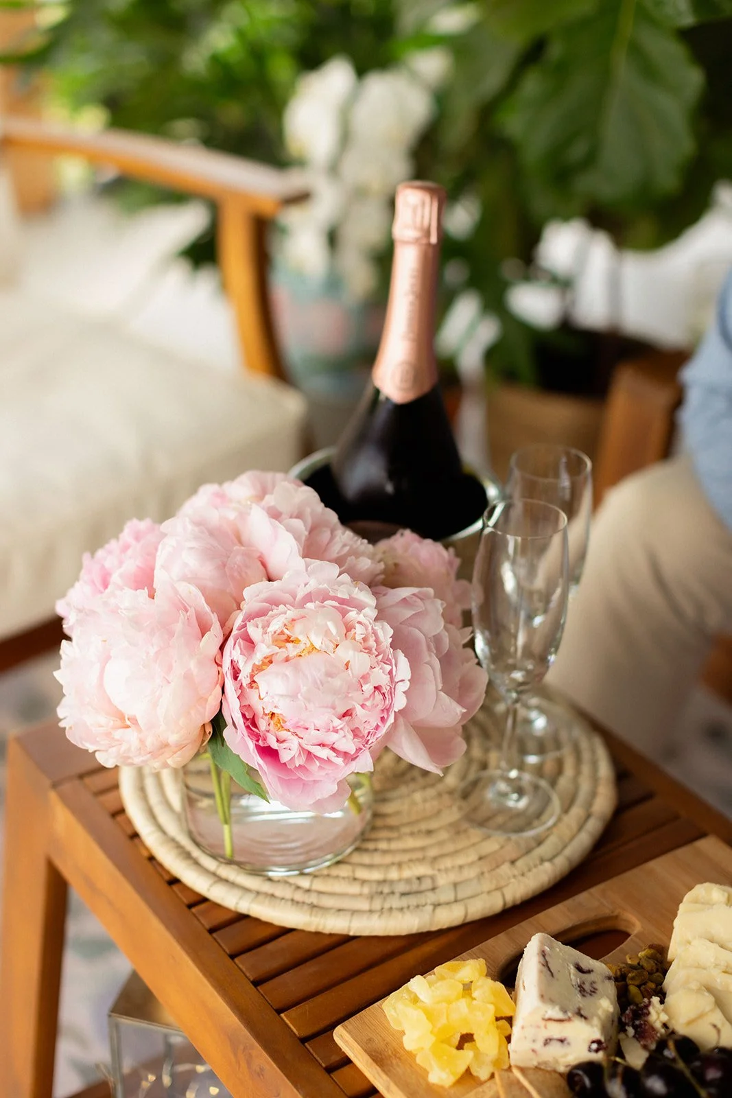 Pink peony flowers in a glass jar on a wooden table next to a bottle of champagne and two empty champagne flutes, with cheese and dried fruit nearby.