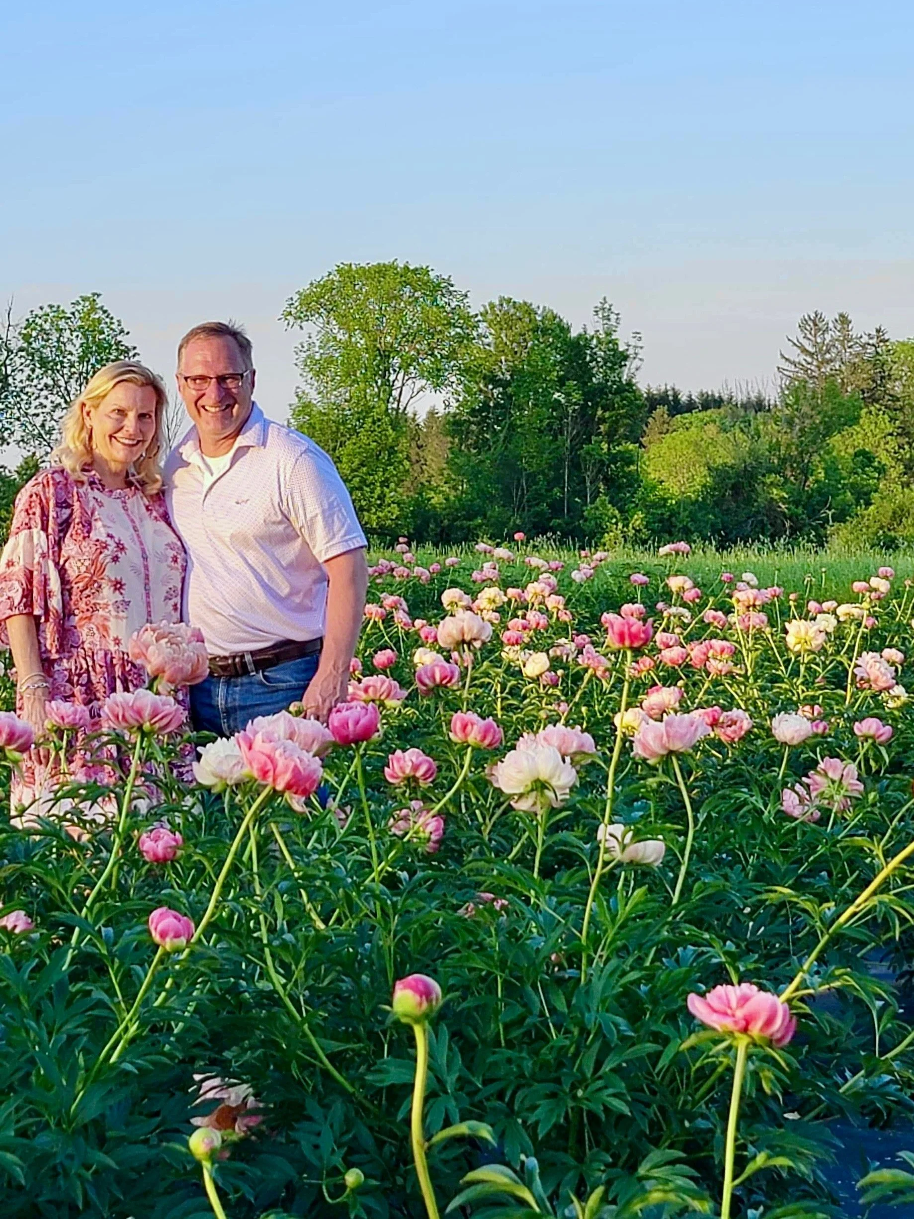 A smiling man and woman standing together in a blooming flower field with tall green trees and a clear blue sky in the background.