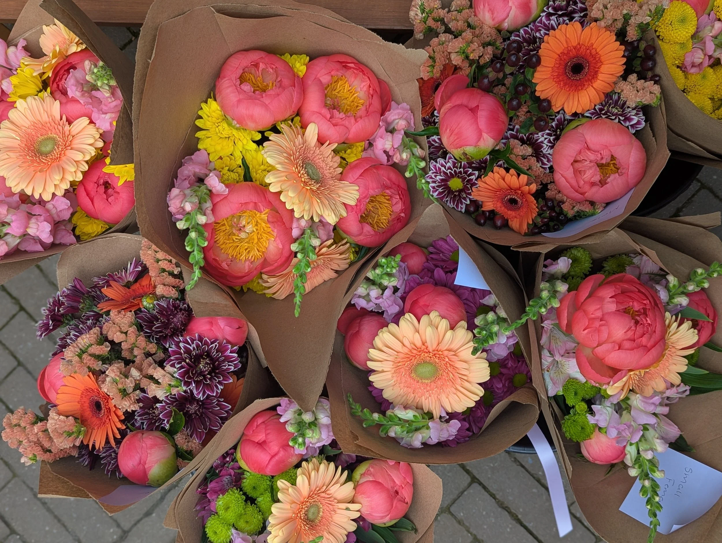 Several bouquets of colorful flowers wrapped in brown paper, including pink peonies, orange gerbera daisies, purple and pink chrysanthemums, yellow chrysanthemums, and small green accents, arranged on a stone sidewalk.