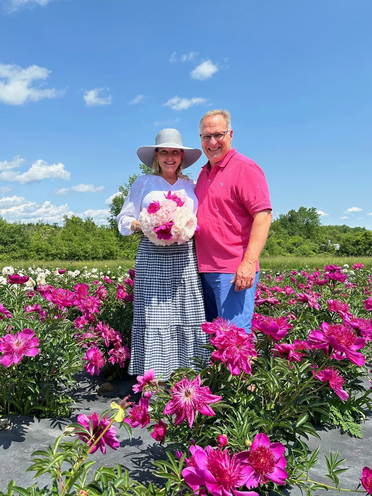 A smiling couple standing in a field of pink flowers under a blue sky with scattered clouds. The woman is wearing a large sunhat, white blouse, and a checkered skirt, holding a bouquet of pink and white flowers. The man is wearing glasses, a pink polo shirt, and blue pants.