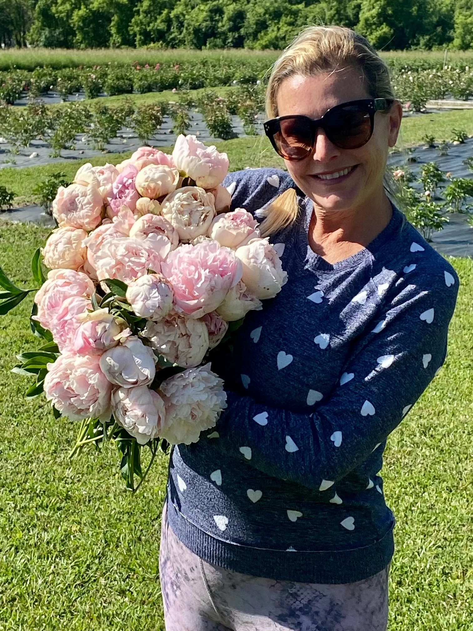 Woman with blonde hair, wearing sunglasses and a navy blue sweater with white hearts, holding a large bouquet of pink and white peonies in a field of blooming peony plants.