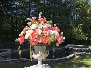 A large floral arrangement in a white urn, featuring pink and white flowers, set outdoors near a fountain with trees in the background.