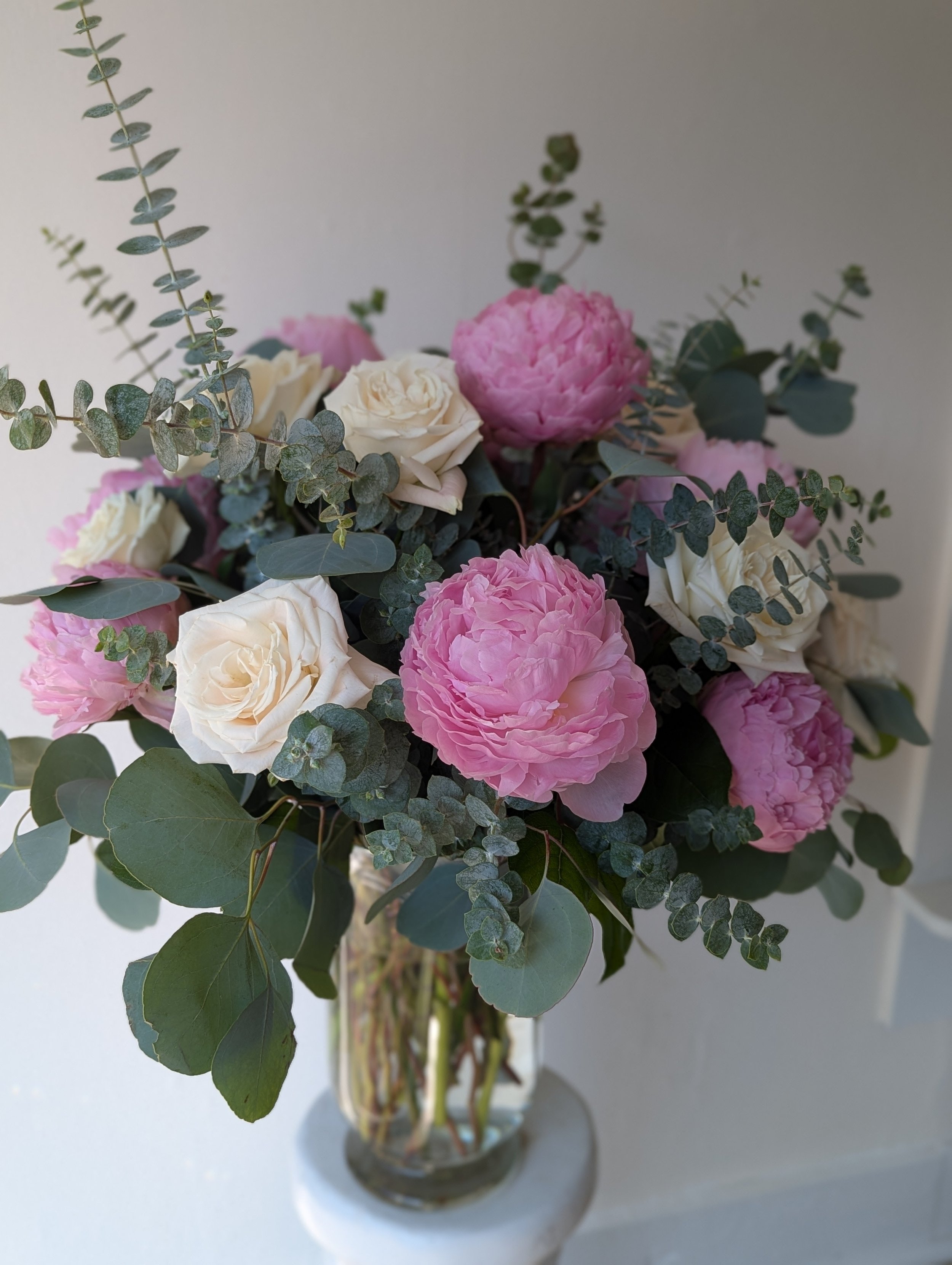 A floral arrangement in a glass vase with pink peonies, white roses, and green eucalyptus leaves.