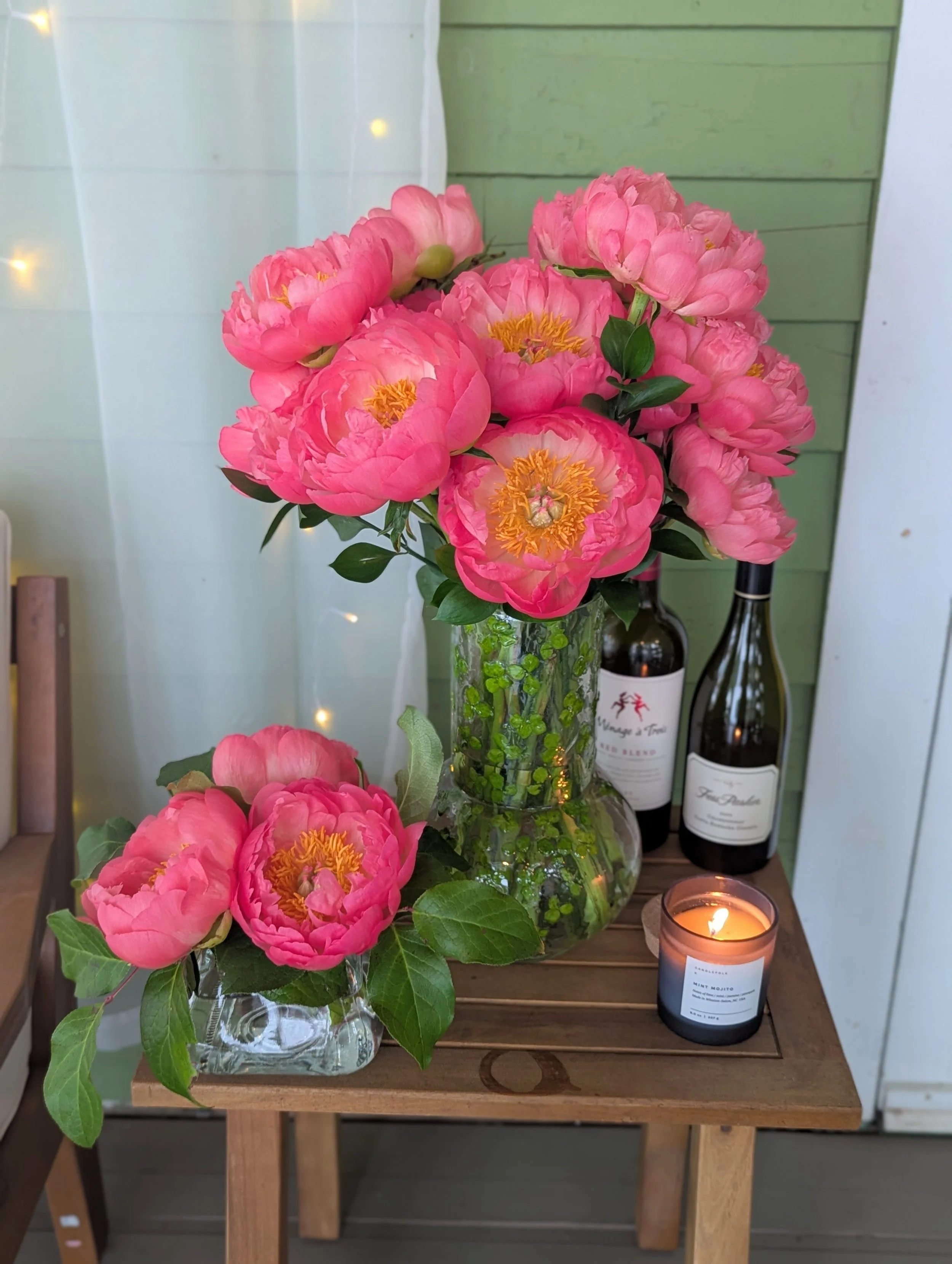 Pink peony flowers in a glass vase with green leaves, placing on a wooden table, with a smaller arrangement of peonies, a lit candle, and two bottles of wine in the background.