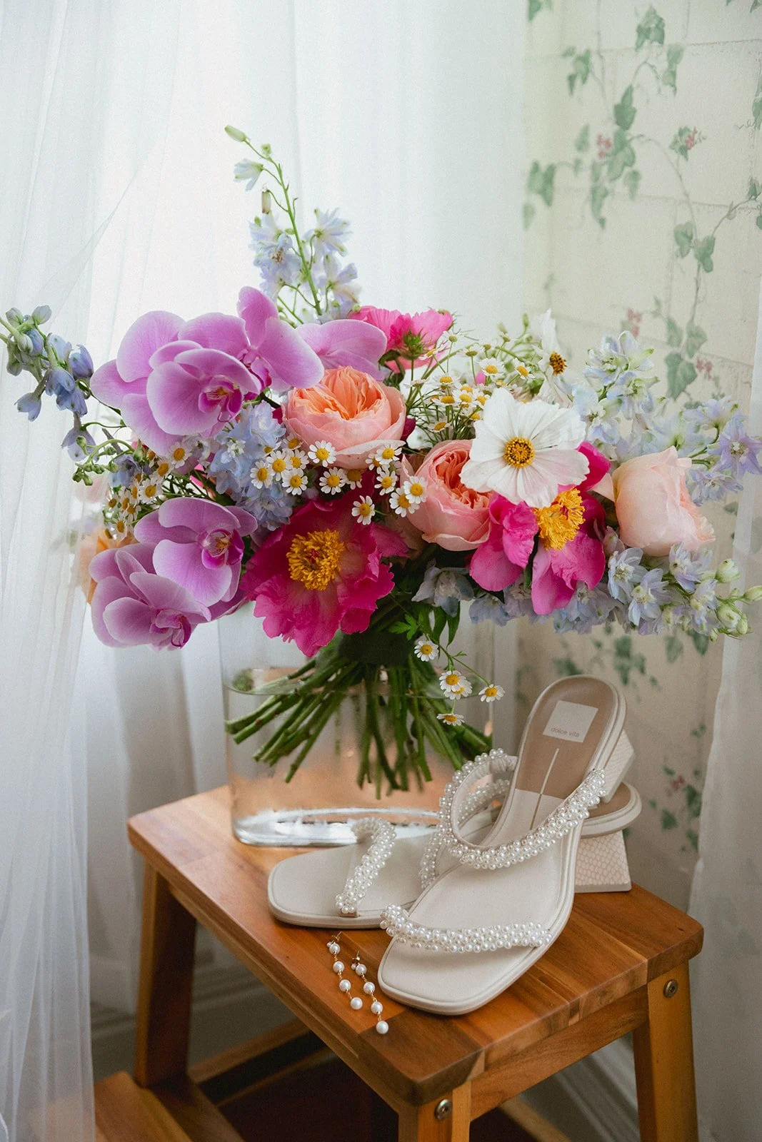 A large bouquet of assorted pink, purple, white, and yellow flowers in a glass vase on a wooden table, with a pair of white beaded high-heeled sandals and pearl jewelry.