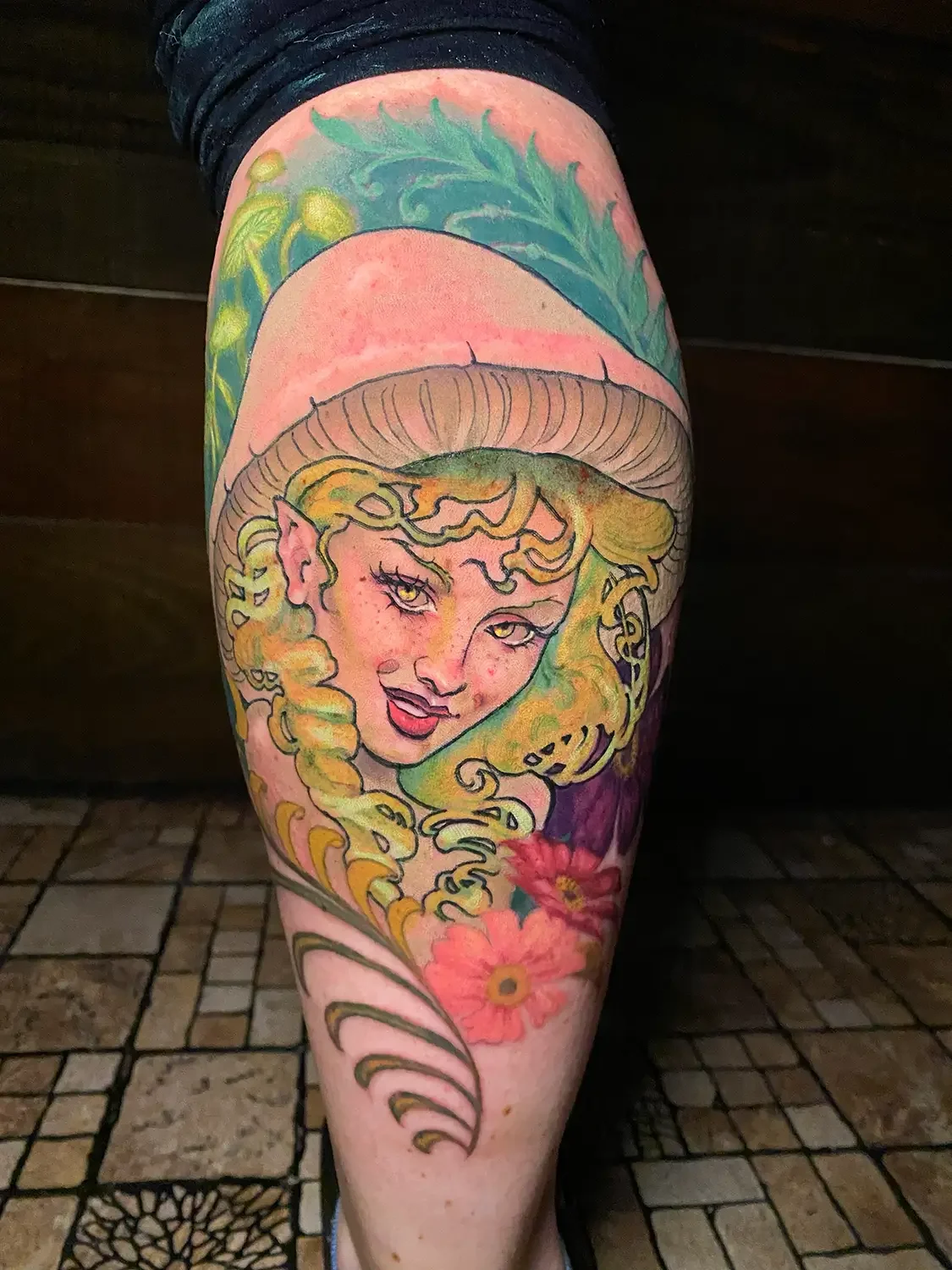 Fantasy color tattoo of a woman with curly hair, a mushroom cap hat, and flowers beneath. Custom Design by RVA tattoo artist Fronkie.