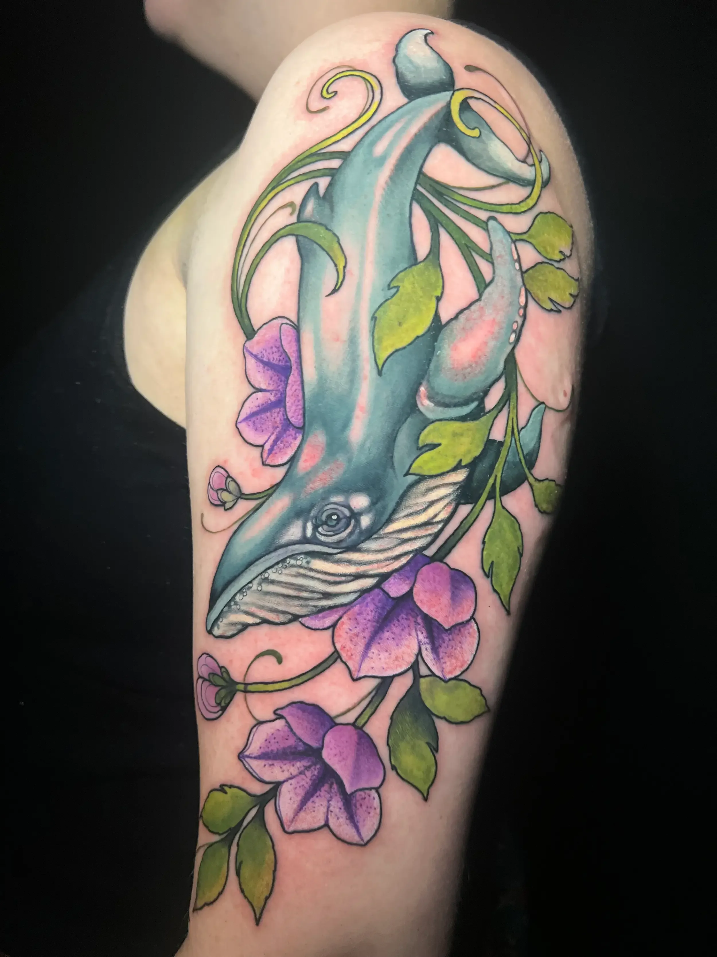 Colorful tattoo of a whale surrounded by purple flowers and green leaves based on A Hitchhiker's Guide to the Galaxy on a person's arm. Done by female tattoo artist Dana Carlson in Virginia.