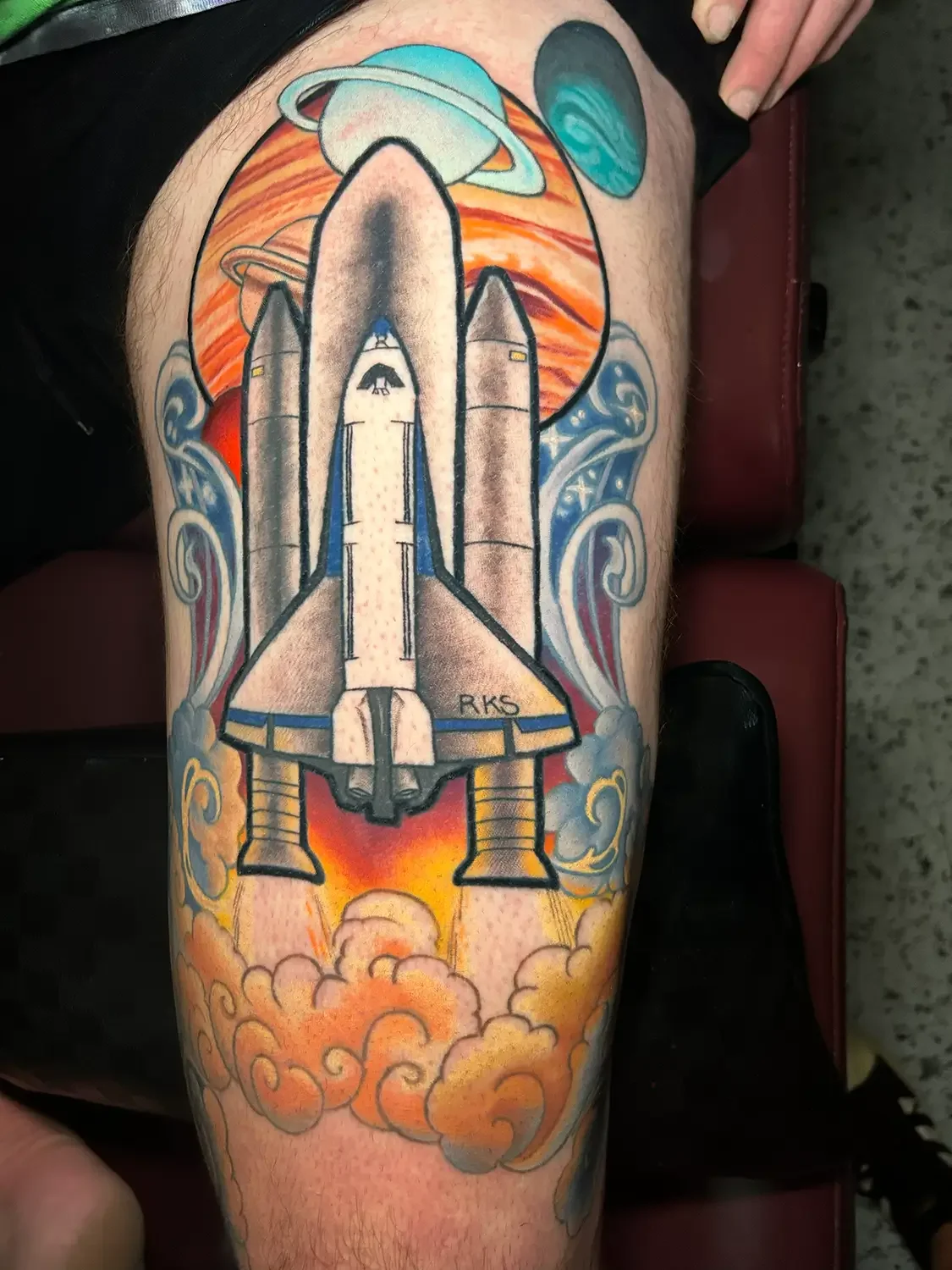 A colorful thigh tattoo of a space shuttle launching with flames and smoke, surrounded by planets and planets with rings in space background. Custom Design by RVA tattoo artist Fronkie.