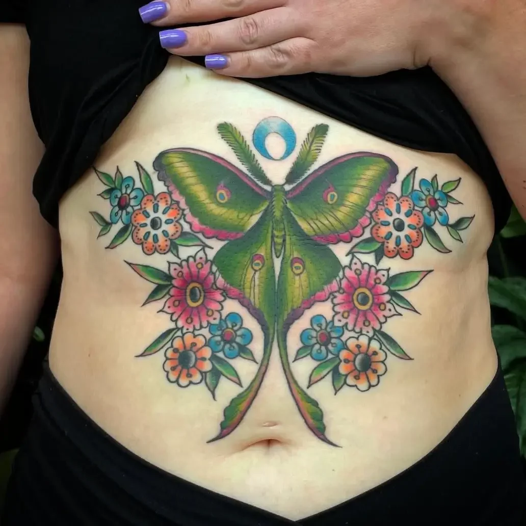 Colorful tattoo of a large green moth with pink and purple accents, surrounded by orange and blue flowers on a person's lower abdomen. Custom design by Jess, tattoo artist in Richmond, Virginia.