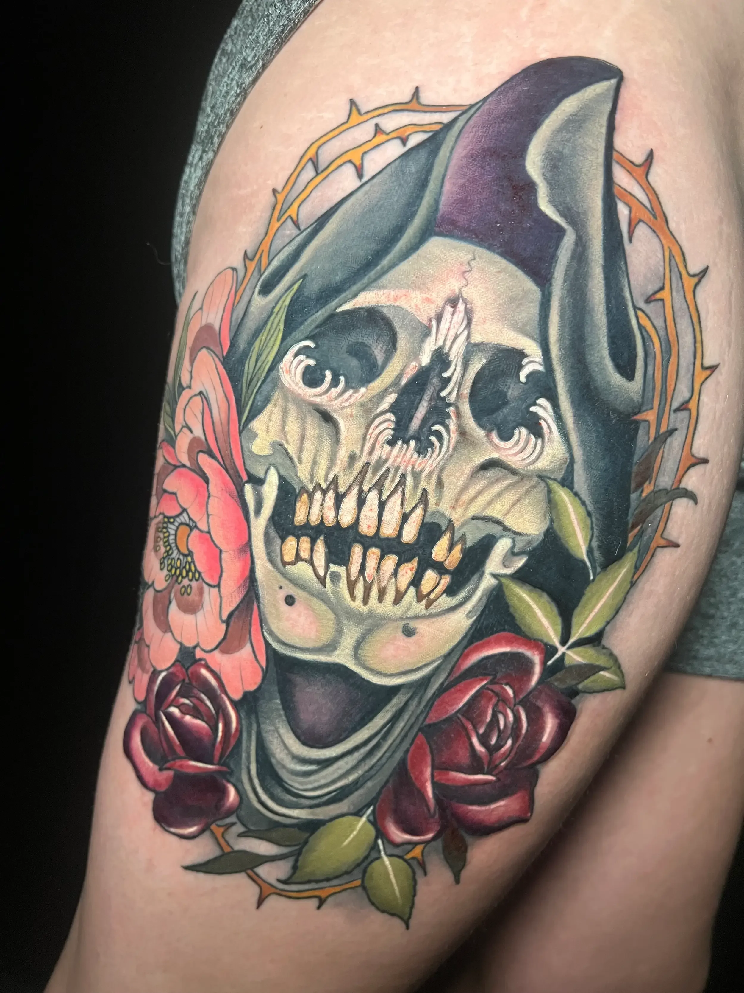 Neo-trad tattoo of a skull wearing a dark hood, surrounded by pink and red flowers with green leaves, and an orange thorny circular border. Custom Design by female tattoo artist Dana Carlson in RVA.