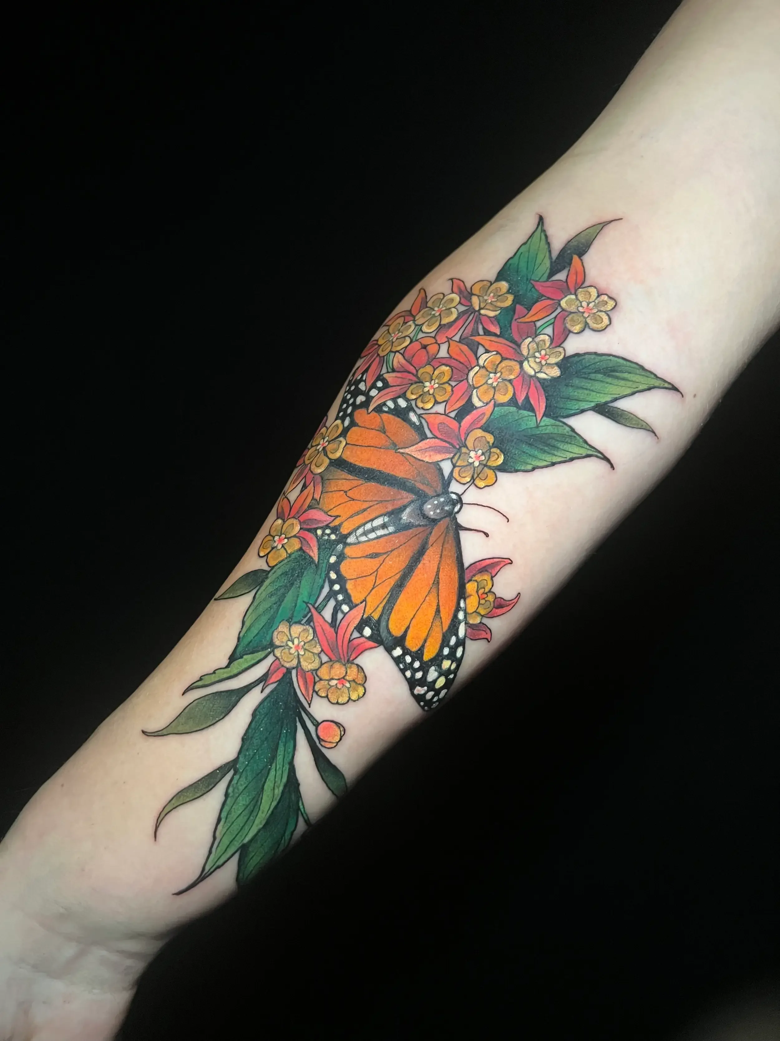 Colorful tattoo of a Monarch butterfly surrounded by green leaves and yellow and red flowers on a person's forearm. Custom Design by female tattoo artist Dana Carlson in RVA.