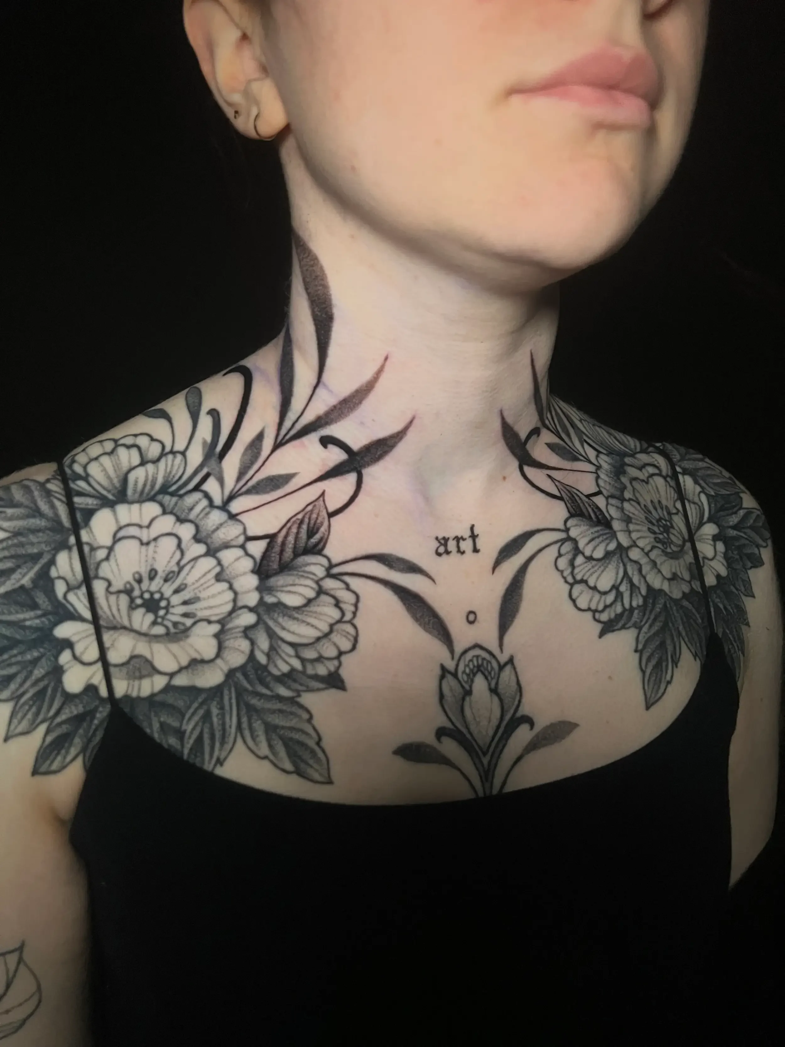 A woman with black and gray floral tattoo on her chest and neck, with the word 'art' in small letters on her chest, wearing a black top, against a black background.