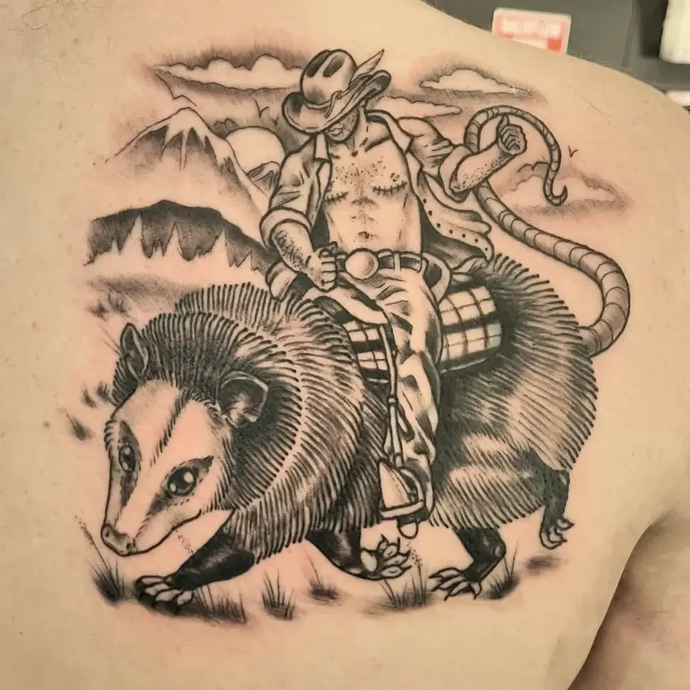 Black and gray tattoo of a cowboy riding a opossum, with mountains and clouds in the background. Custom design by Jess, tattoo artist in Richmond, Virginia.