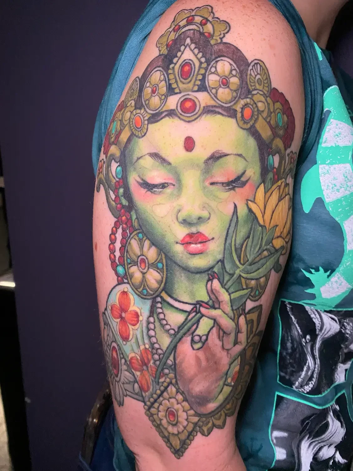 A colorful fantasy tattoo of a green Tara Mantra with closed eyes and elaborate jewelry, holding a flower stem with leaves, on a person's upper arm. Custom Design by RVA tattoo artist Fronkie.