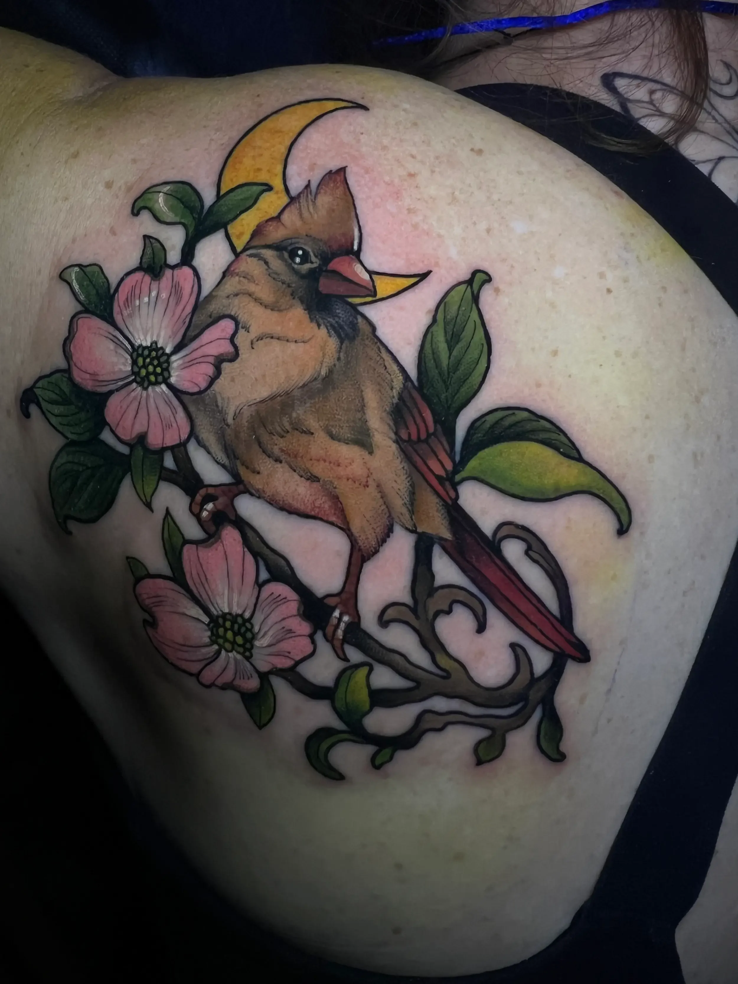 Colorful tattoo of a female cardinal perched on a branch with pink dogwood flowers, green leaves, and a yellow crescent moon in the background. Custom Design by female tattoo artist Dana Carlson in RVA.