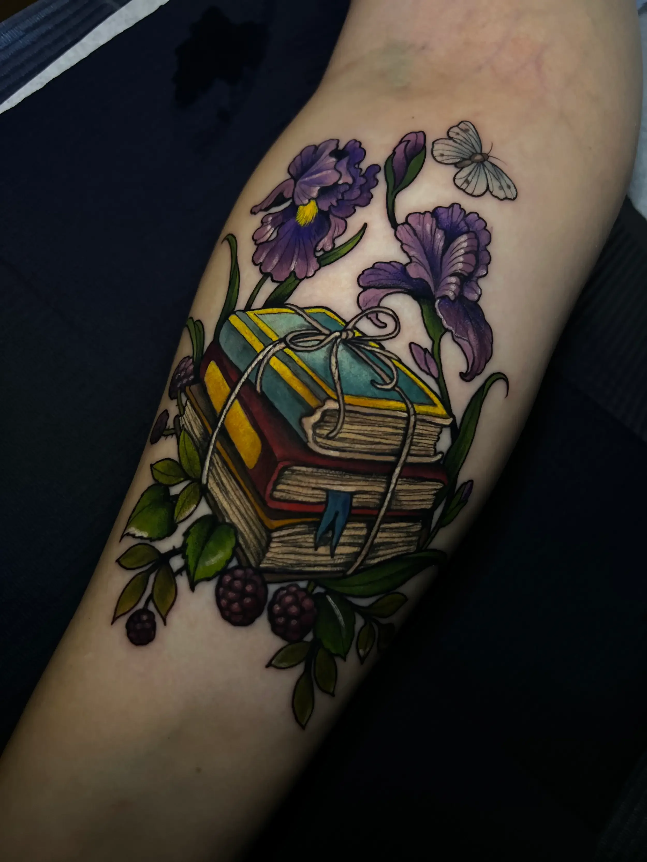 Colorful neo-trad tattoo of stacked books wrapped with string, surrounded by purple irises, green leaves, blackberries, and a small butterfly on the upper right. Custom Design by female tattoo artist Dana Carlson in RVA.