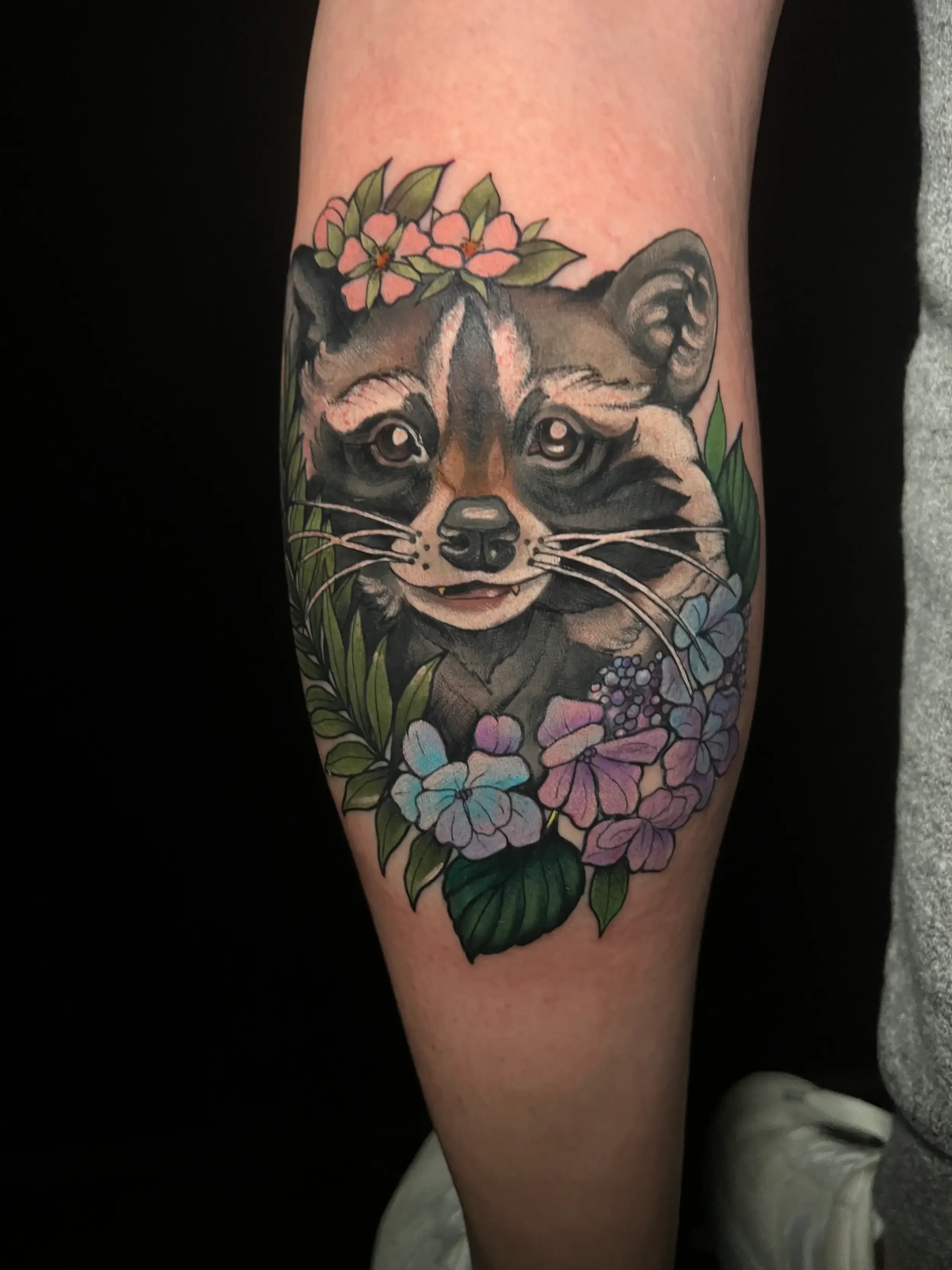 Tattoo of a raccoon surrounded by pink and purple flowers and green leaves on a person's arm. Done by female tattoo artist Dana Carlson in Virginia.
