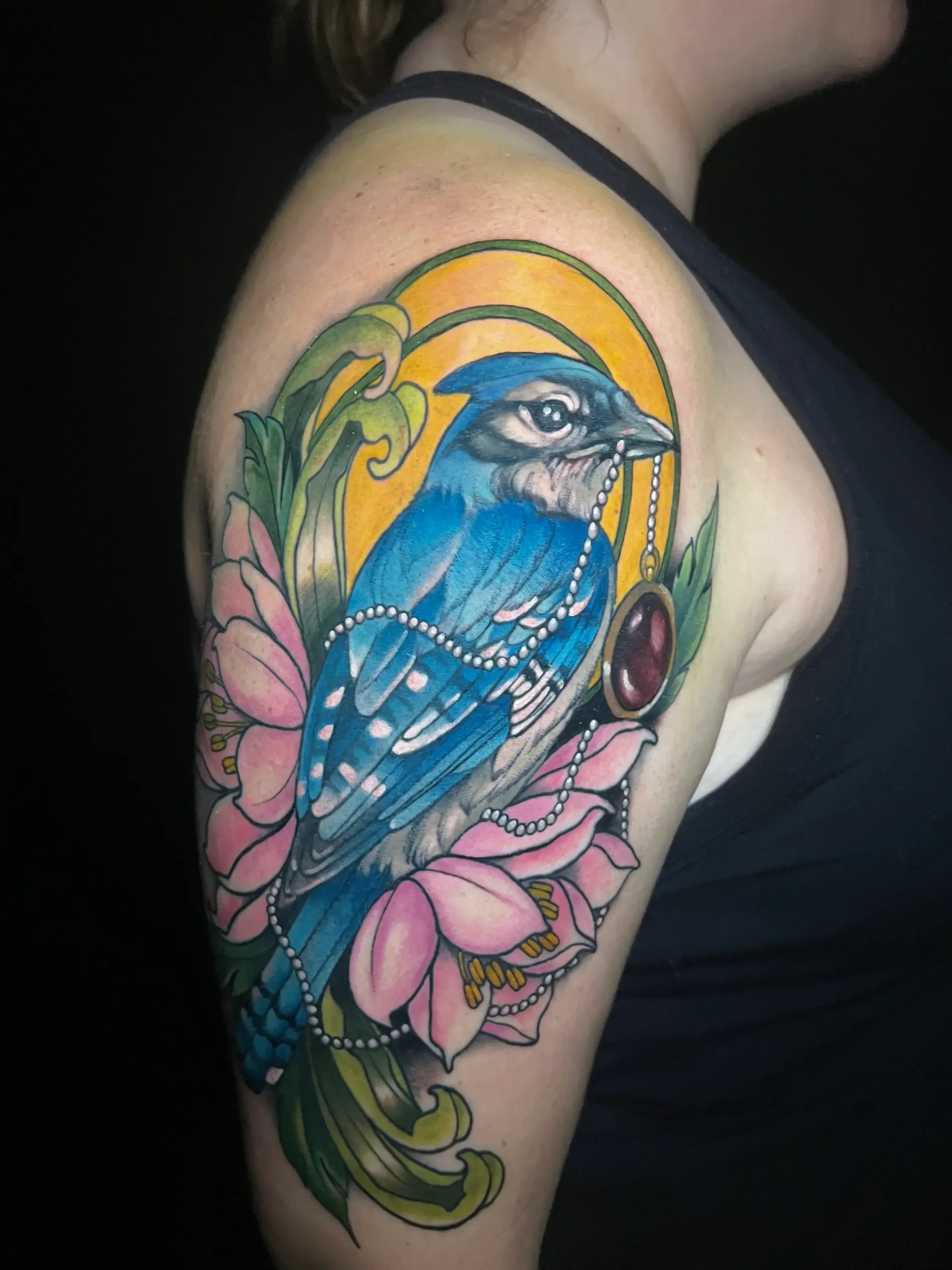 Colorful neo-trad tattoo of a bluebird holding a pearl necklace, surrounded by pink flowers and green foliage, with a yellow background. Custom Design by female tattoo artist Dana Carlson in Virginia Beach.