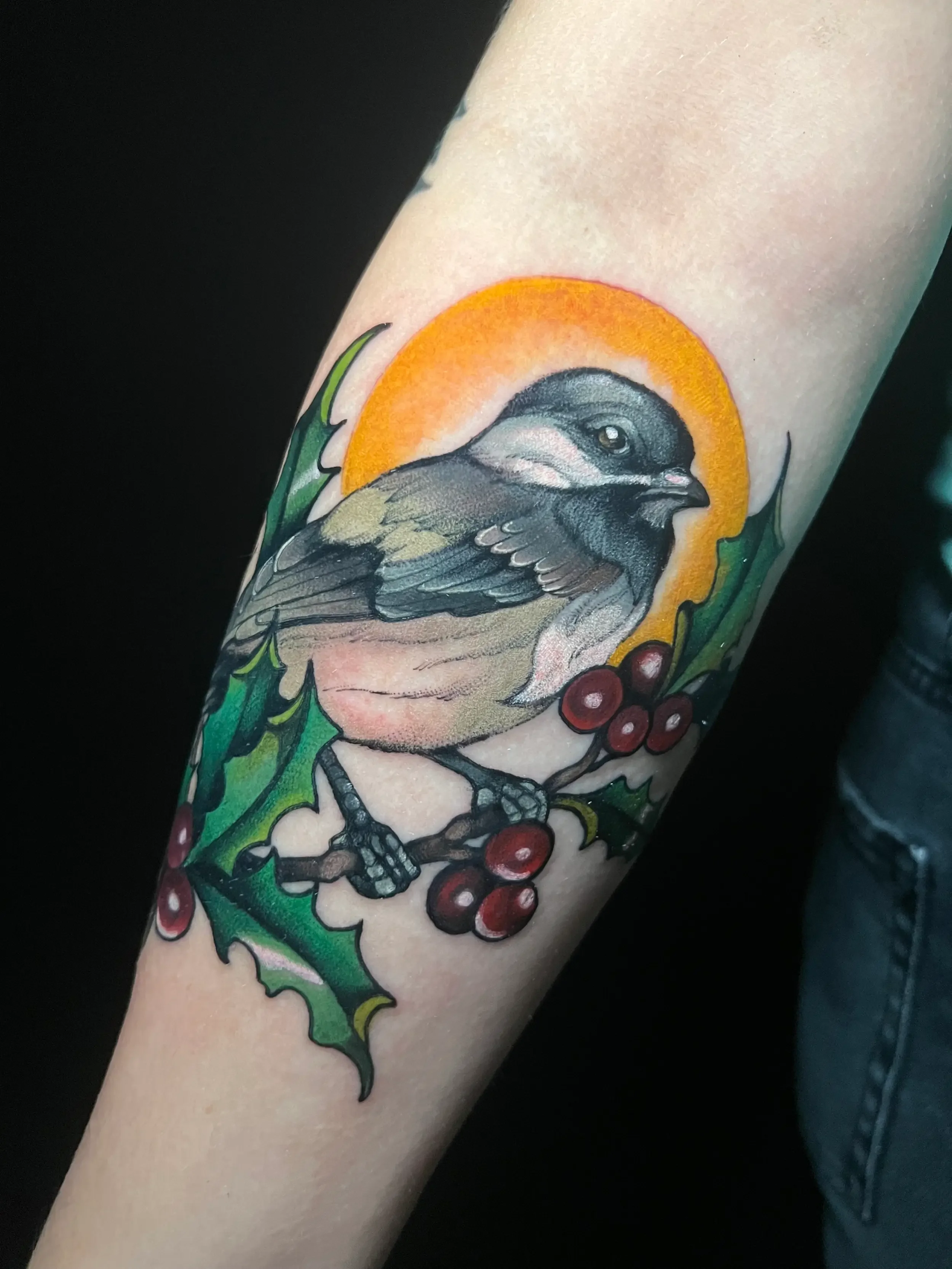 Colorful tattoo of a bird perched on holly leaves and berries, with an orange glow behind. Done by female tattoo artist Dana Carlson in Virginia.