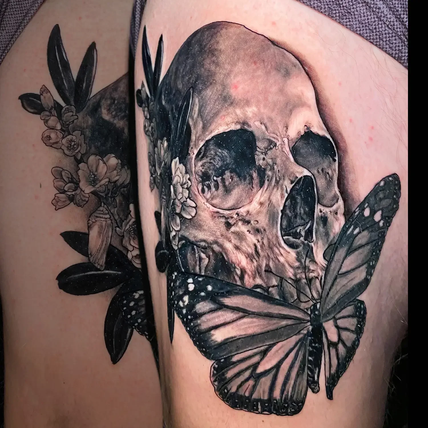 A detailed black and gray tattoo of a human skull with butterflies and flowers surrounding it on a person's leg. Custom Design by RVA tattoo artist Fronkie.
