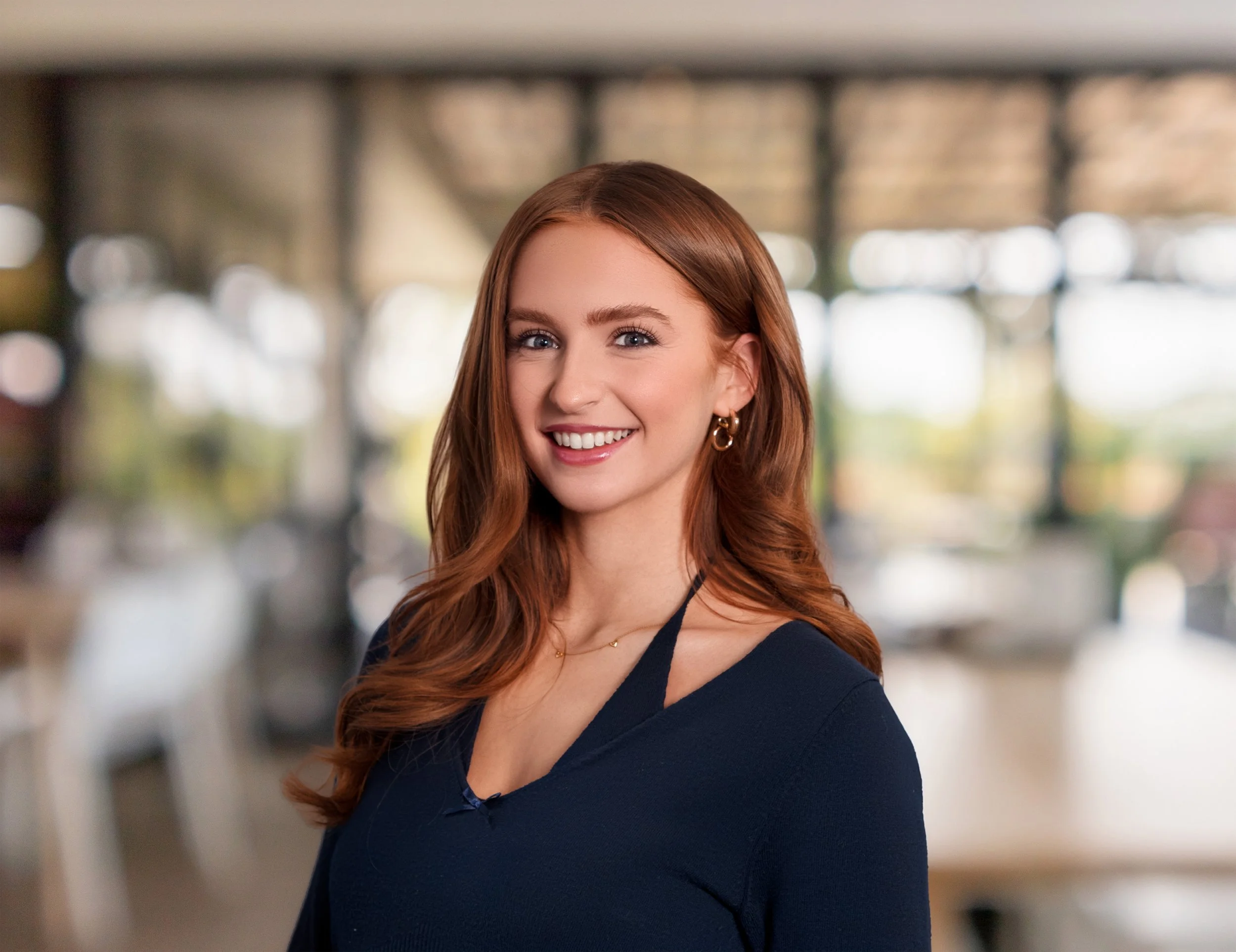 Close-up of a smiling woman with red hair, blue eyes, hoop earrings, and a dark blue top, in an indoor setting with blurry background.