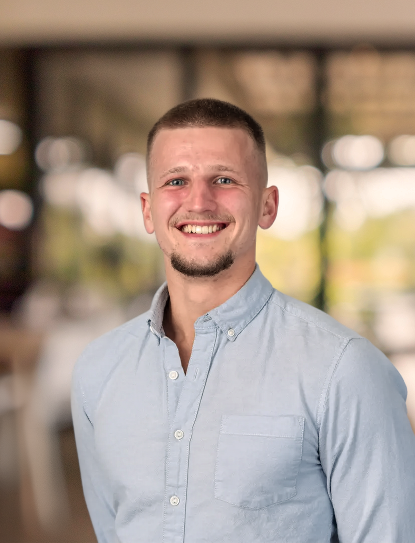 A young man with short light brown hair and a trimmed beard, smiling, wearing a light blue button-up shirt, standing indoors with a blurred background of bright lights and windows.