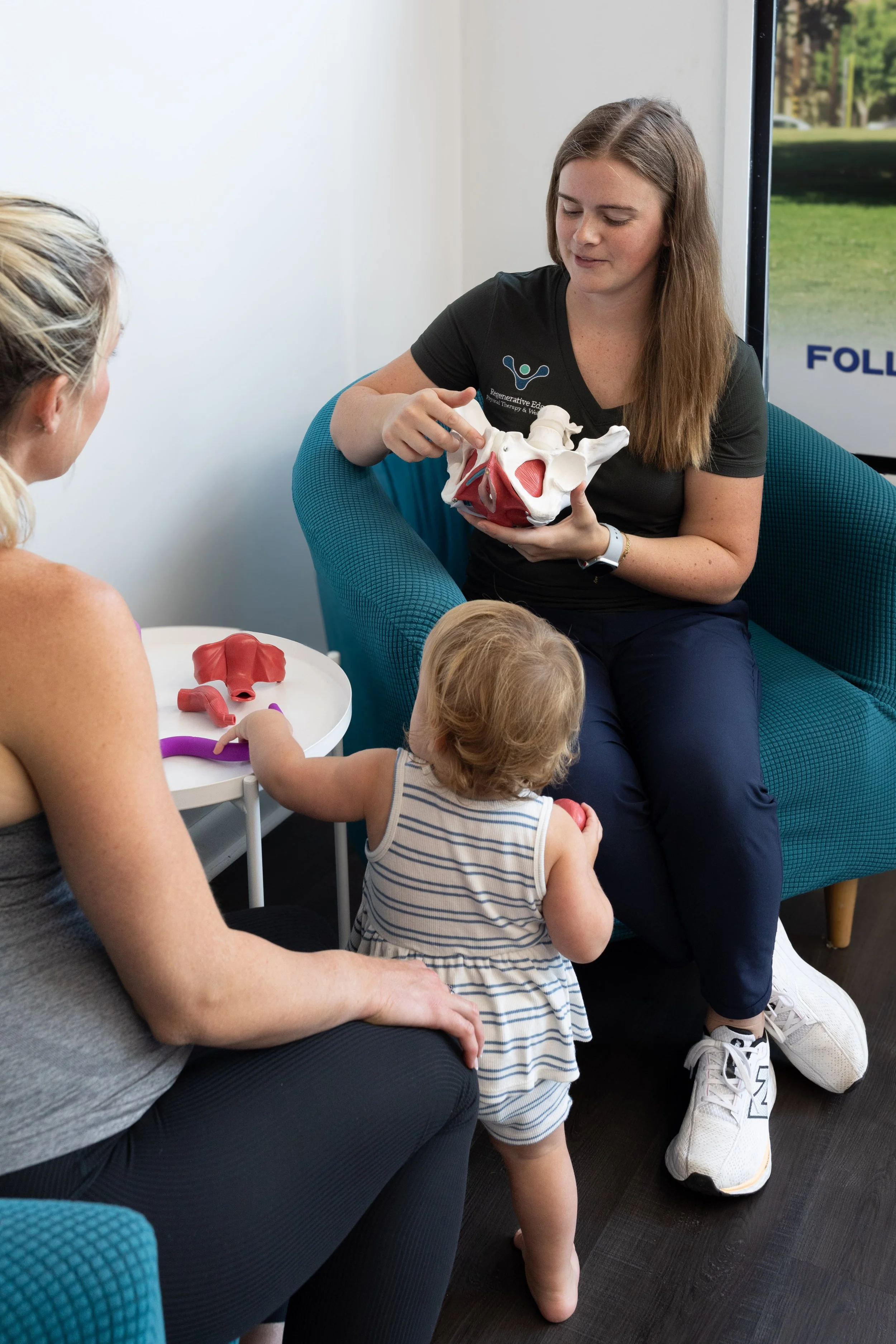 A woman showing a pelvis model to a small child and a woman, with another pelvis model on a small table.