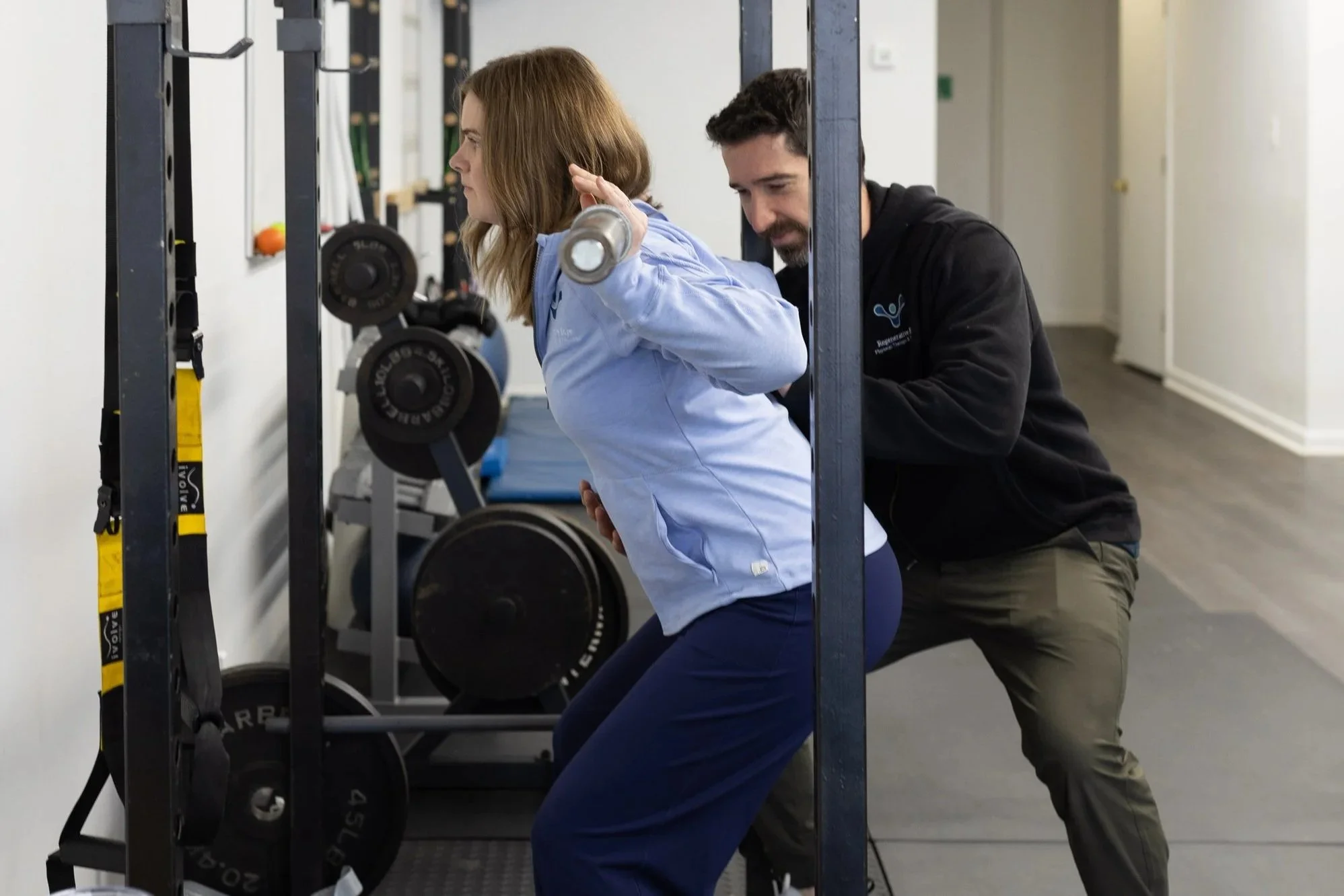 A woman is performing a squat with a barbell on her shoulders while a trainer supports her during a workout in a gym.