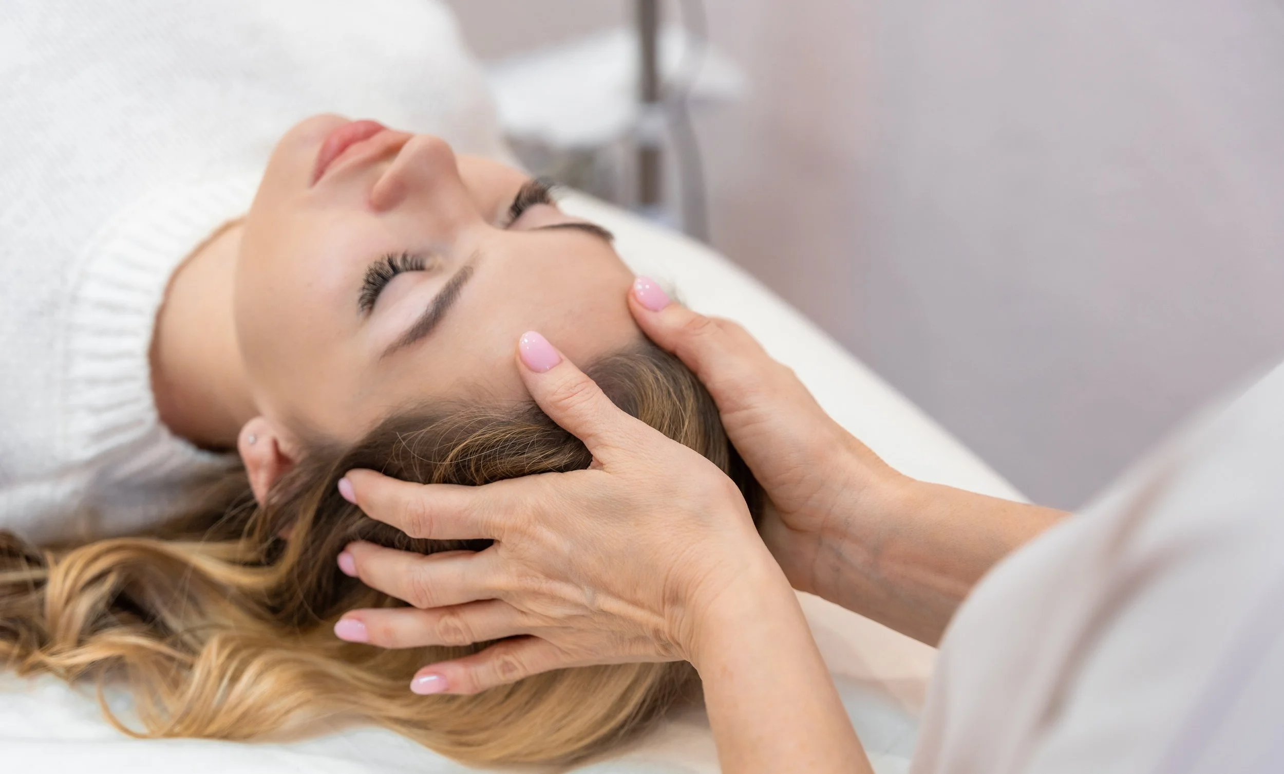 A woman is lying with her eyes closed, receiving a light touch energy work from another person, with hands gently placed on her forehead and temples.