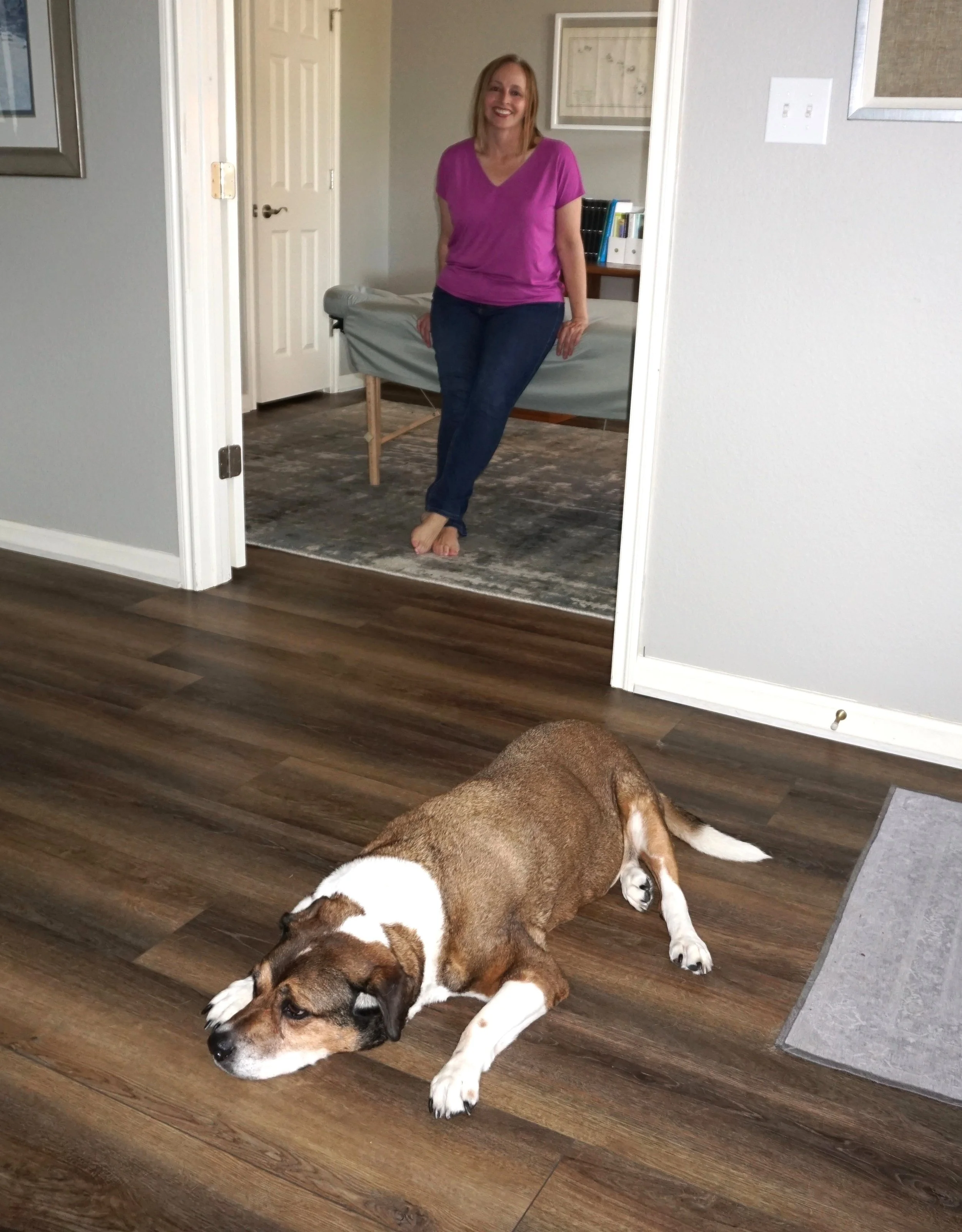 A woman wearing a purple shirt and blue jeans, is smiling and leaning against a doorframe in a room with a dog lying down on a wooden floor.
