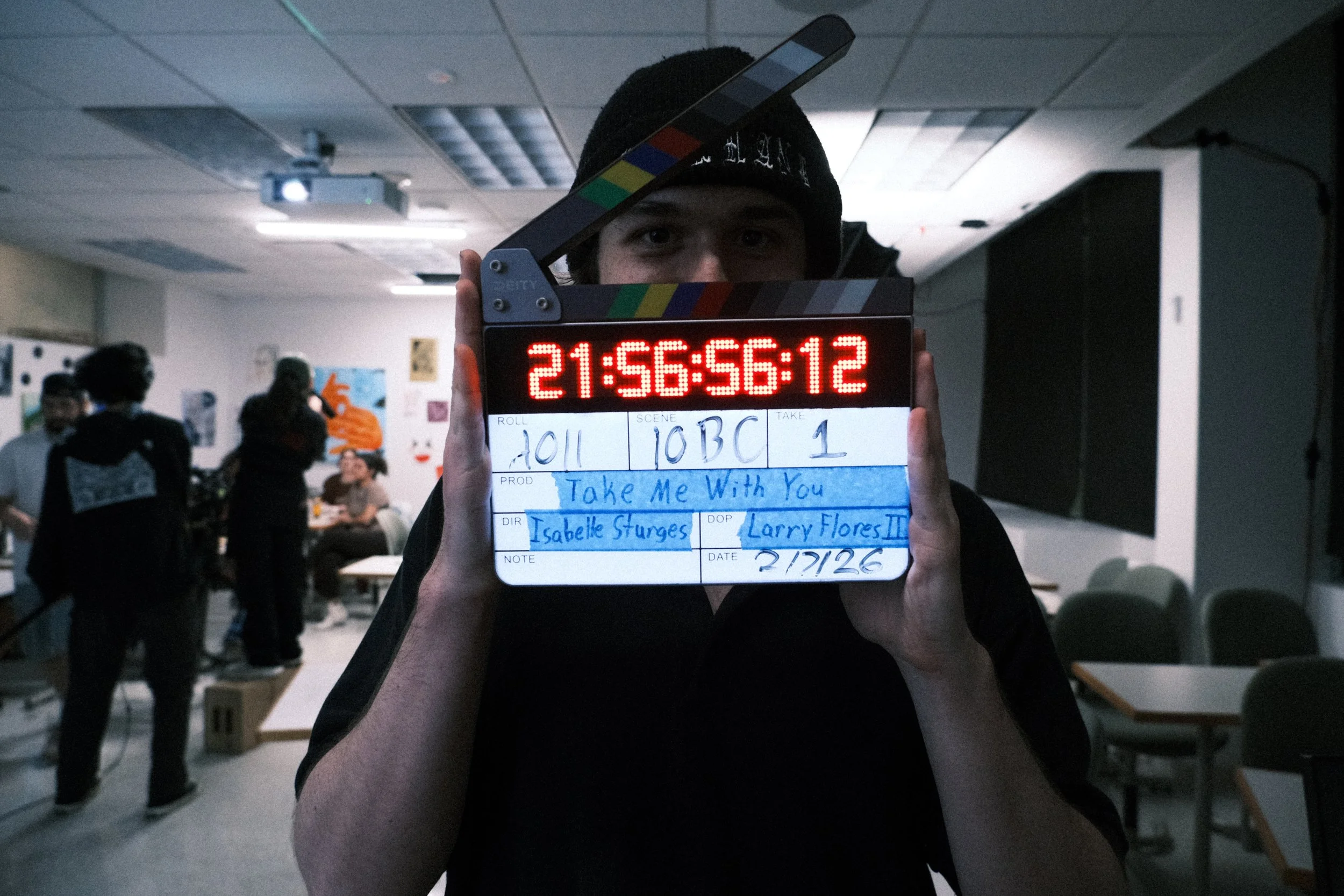 A young man holding a movie clapperboard in a busy indoor setting, with multiple people in the background and ceiling lights overhead.