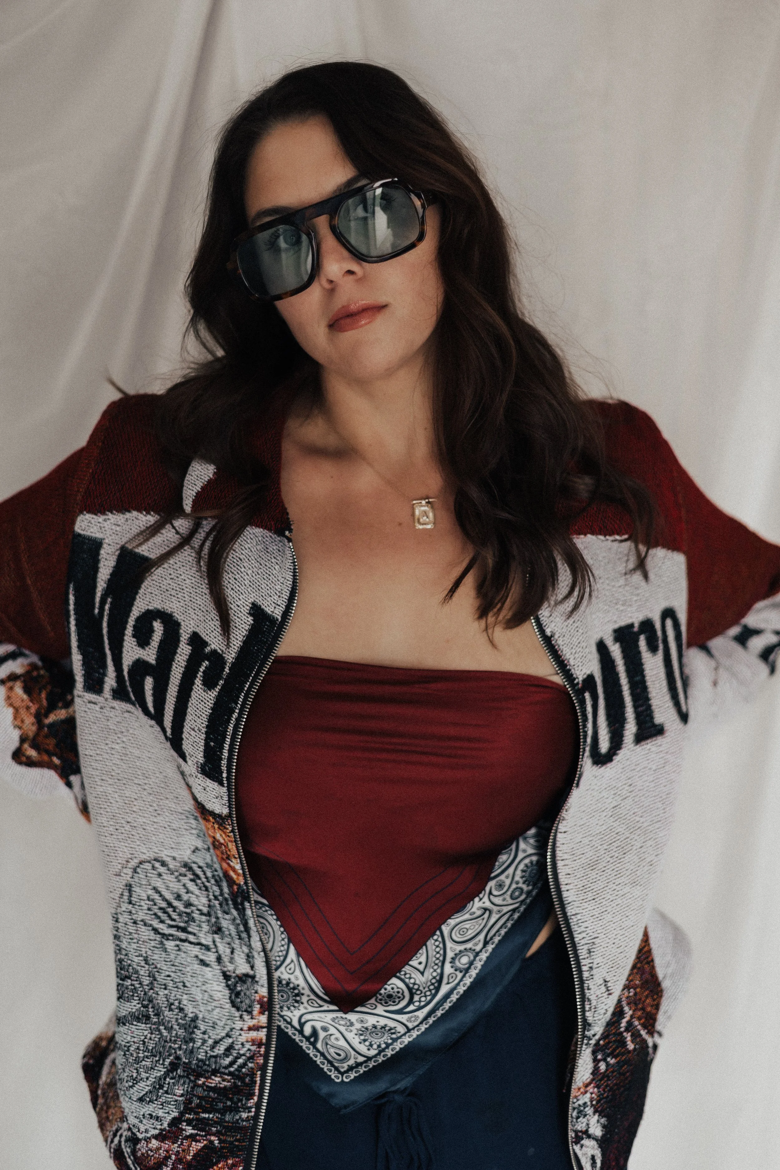 Woman with dark hair wearing large sunglasses, a red top, a patterned bandana, a white jacket with text, and dark pants, standing against a plain background.