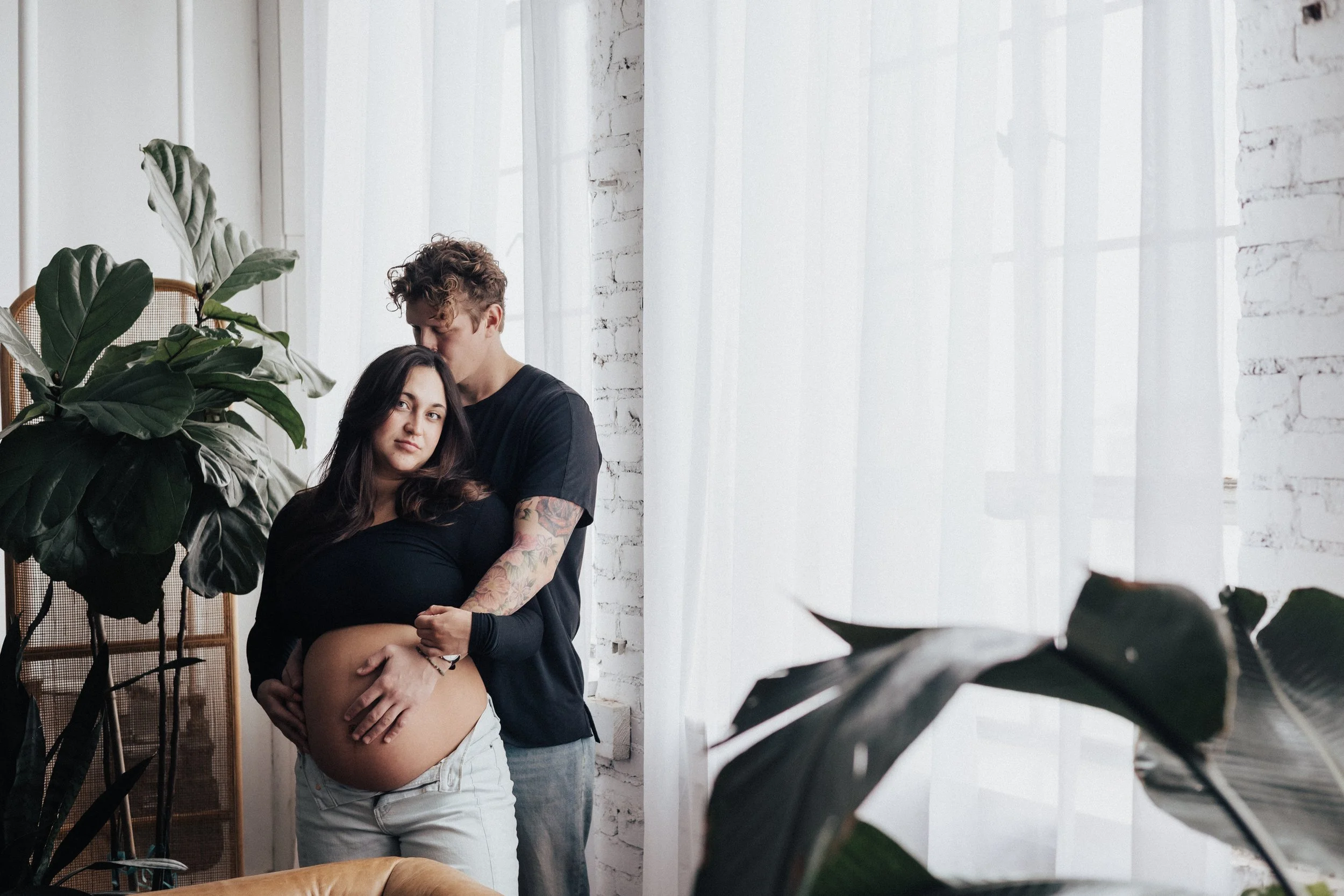 A pregnant woman and her partner standing together near a large window with sheer curtains, with plants in the foreground and a white brick wall background