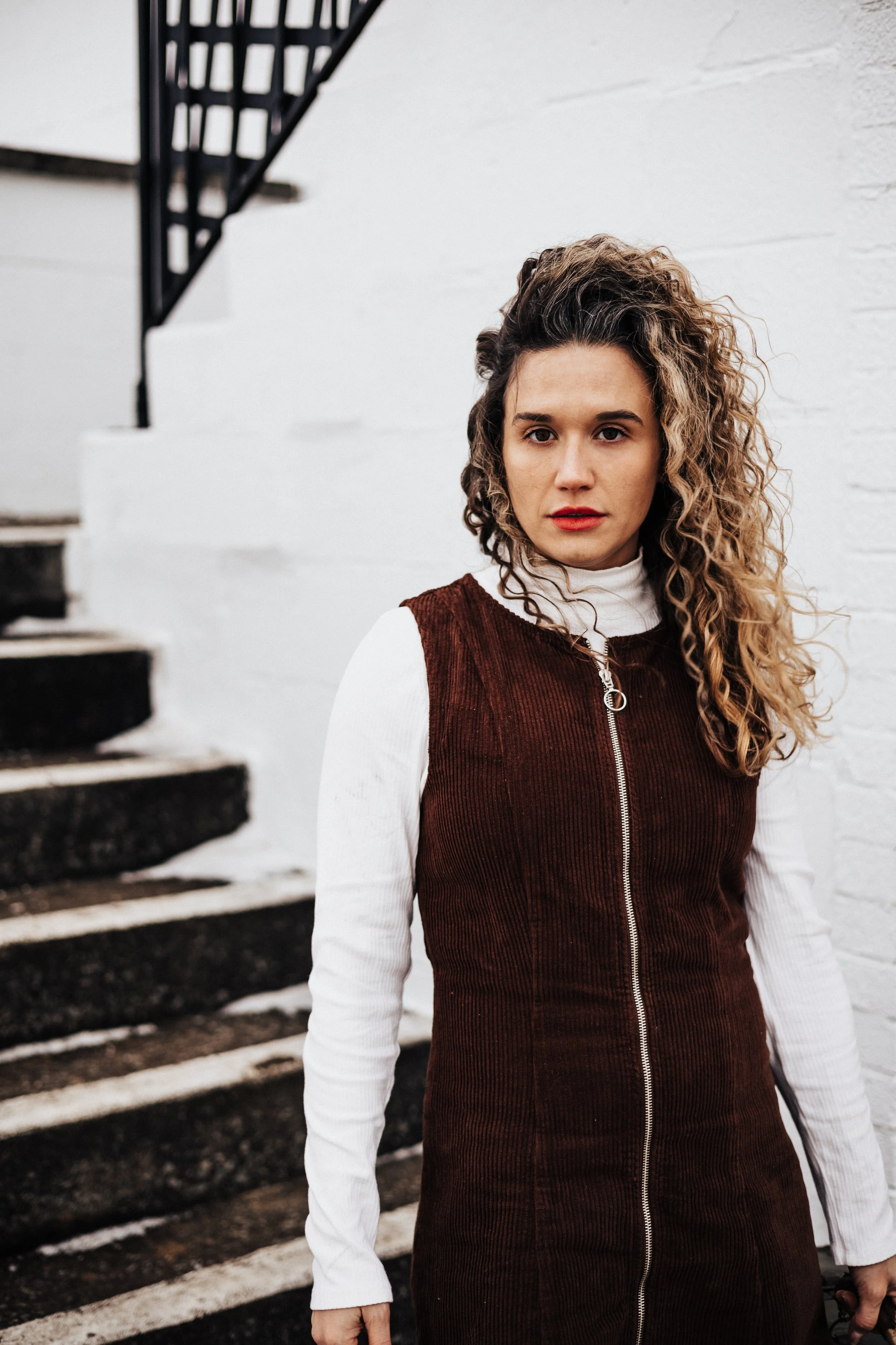 A woman with curly hair wearing a white turtleneck and a brown zip-up sleeveless dress standing next to a staircase against a white brick wall.