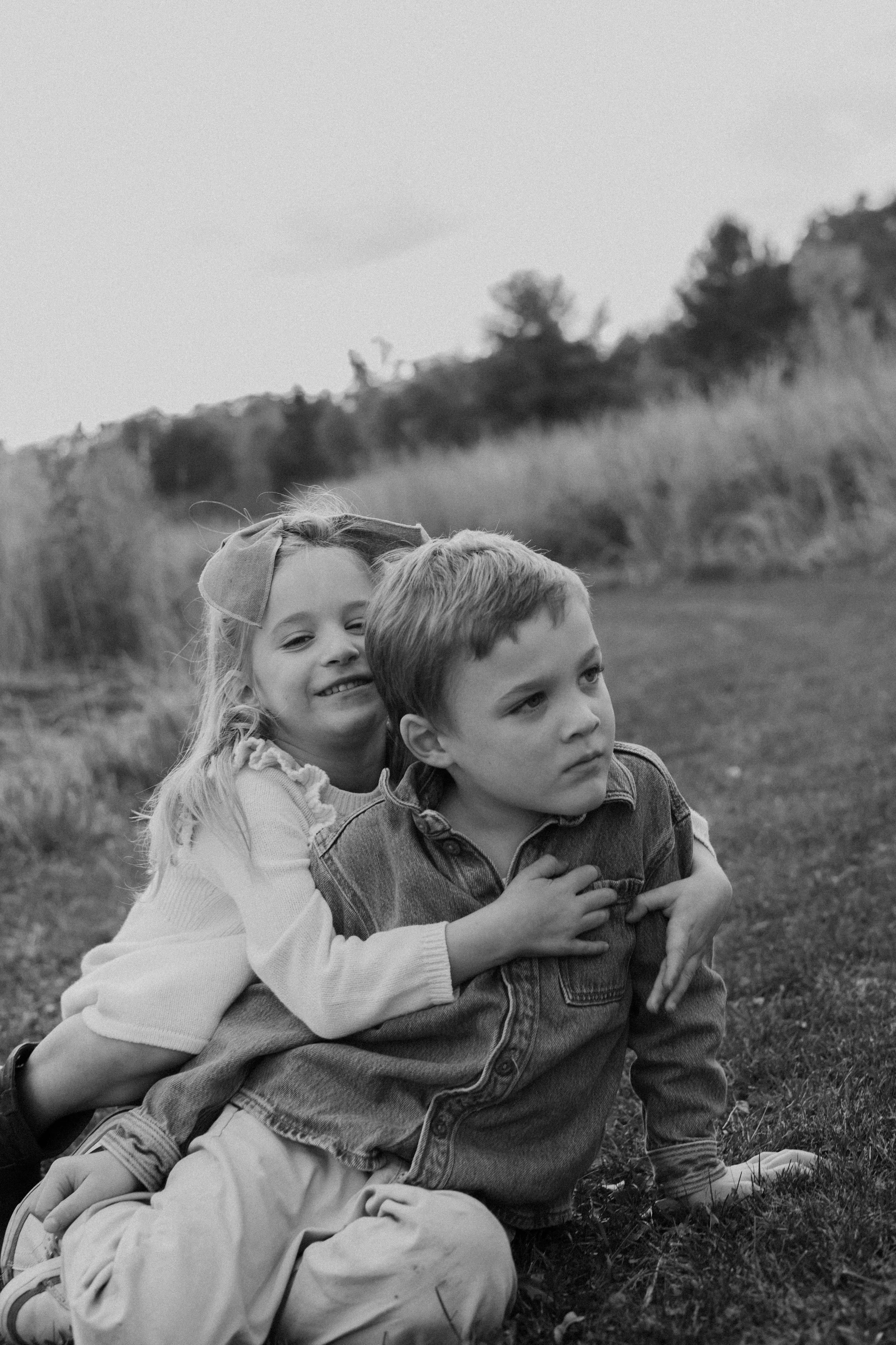 a Calvin Klein inspired Black and white photo of a girl hugging a boy from behind outdoors, with grassy fields and trees in the background.