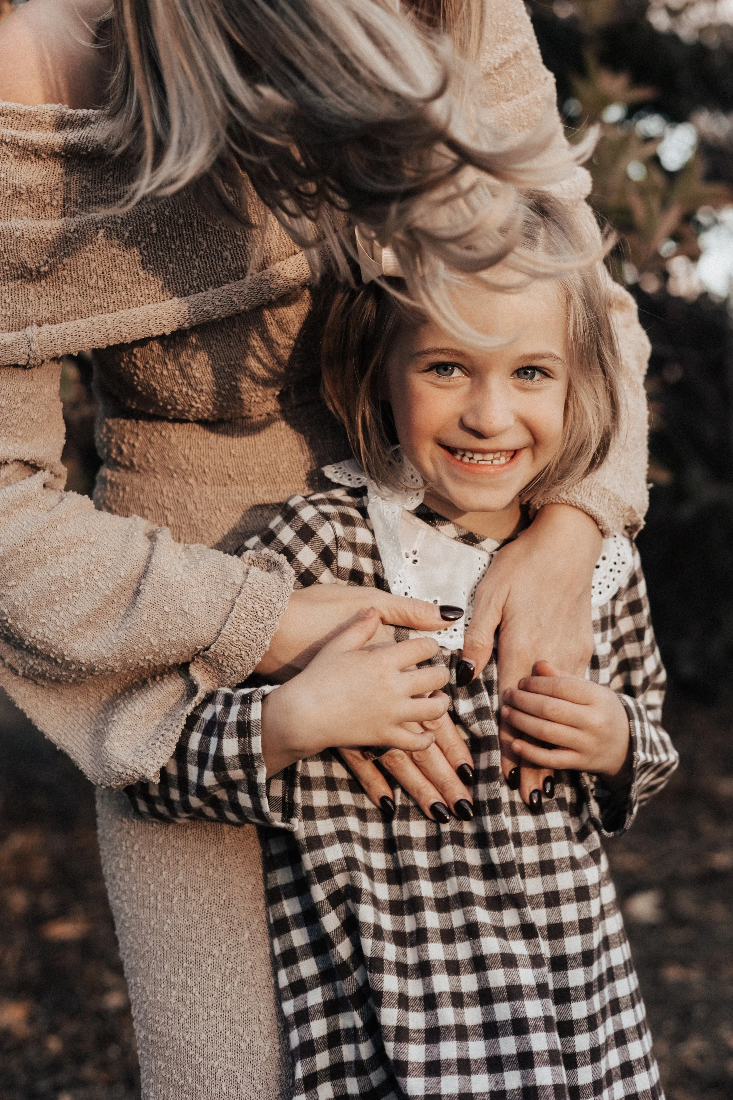 A woman and a young girl smiling outdoors, with the woman hugging the girl from behind, both wearing cozy, warm clothing.