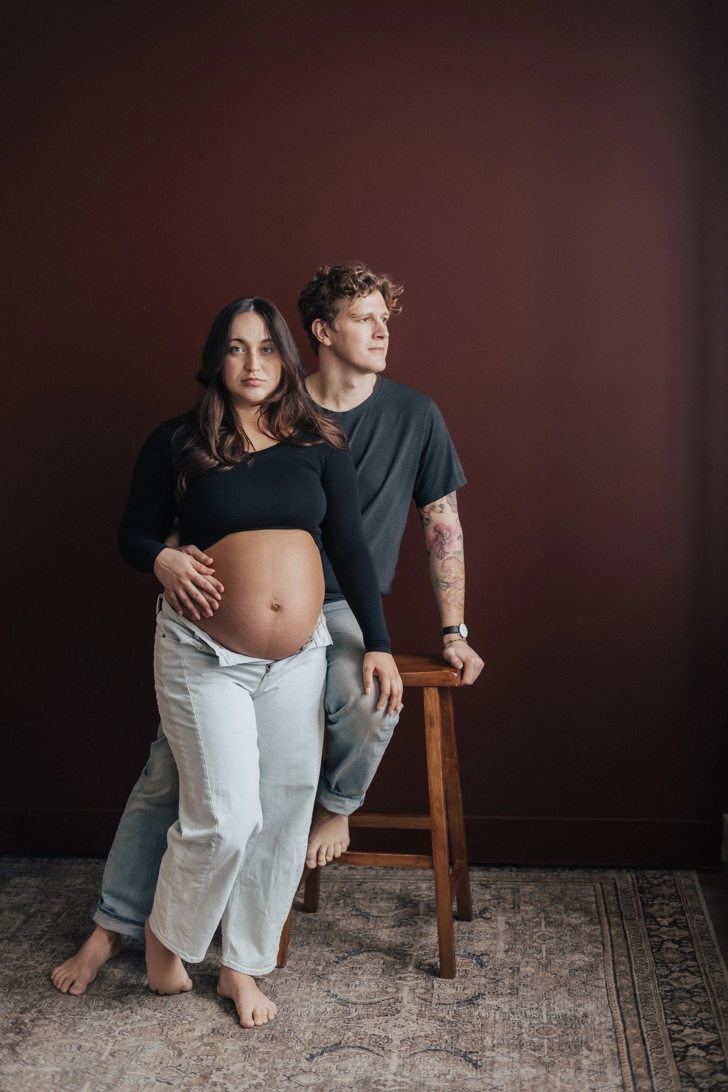 A pregnant woman and a man posing together indoors. The woman is standing barefoot, wearing a black long-sleeve crop top and white pants, with her hand on her belly. The man is sitting on a stool, casually dressed in a dark T-shirt and jeans, with ta