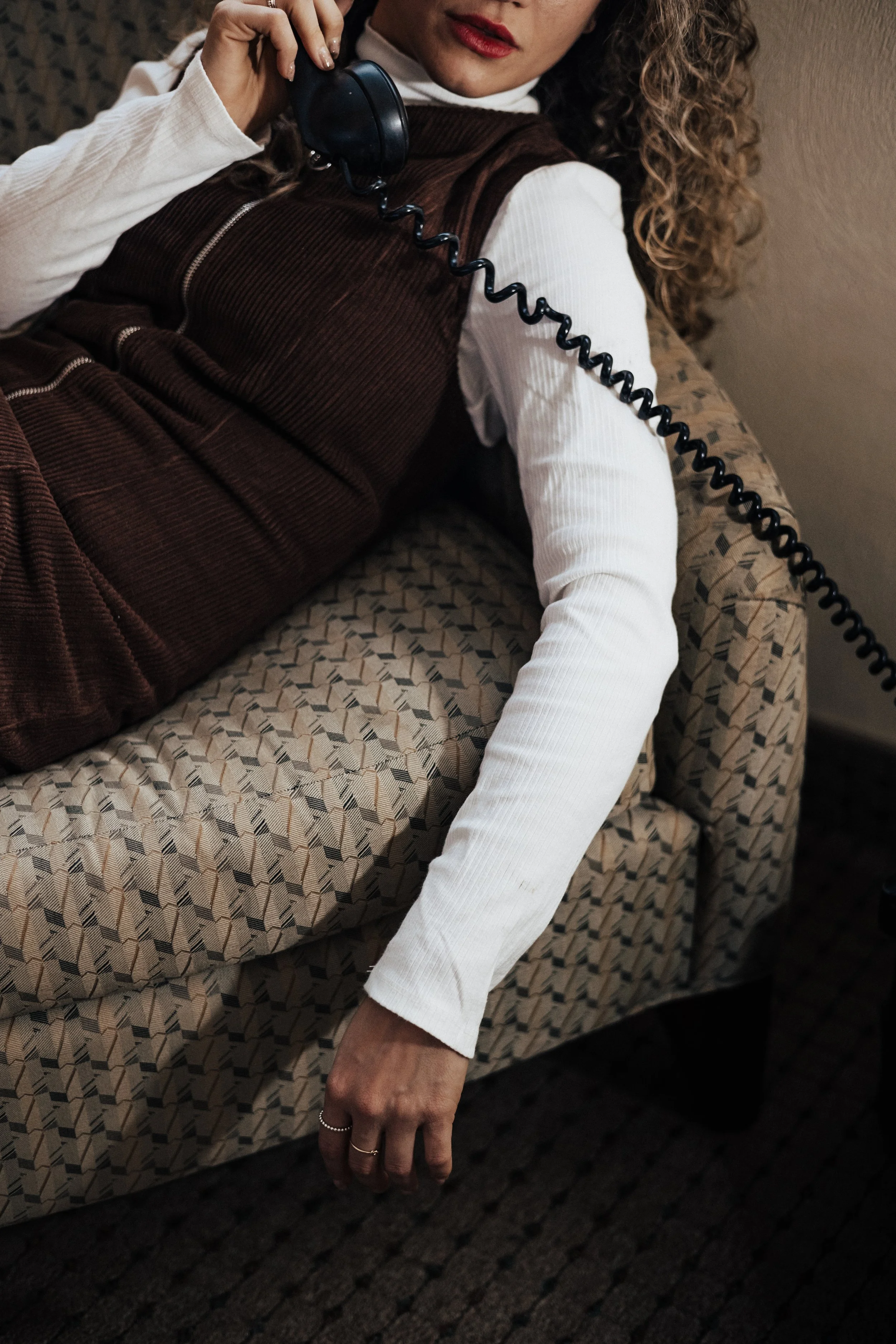 A woman with curly hair, wearing a white long-sleeve shirt and brown corduroy vest, is lying on a patterned sofa while talking on an old-fashioned corded phone for a branding photography shoot in ohio