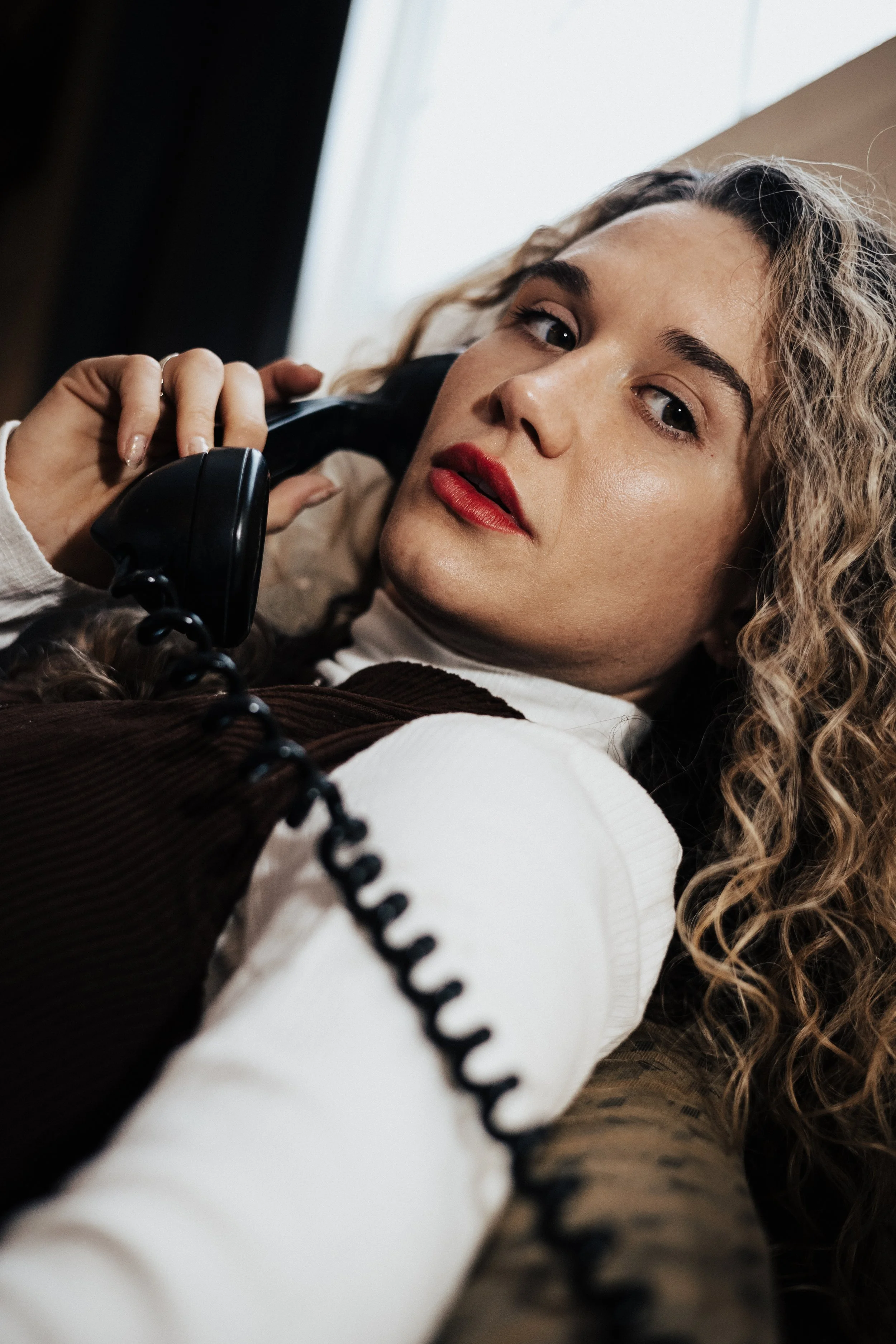 Woman with curly hair and red lipstick talking on a vintage black rotary phone in an old midwest Ohio motel.