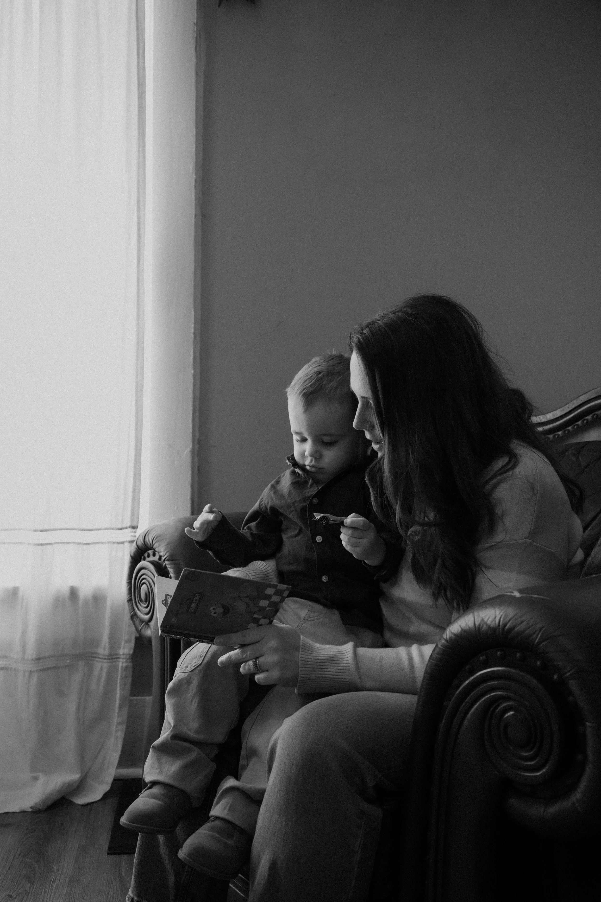 A woman and a young boy sitting together on a vintage leather couch, engaging with a book and a small object, near a window with light curtains