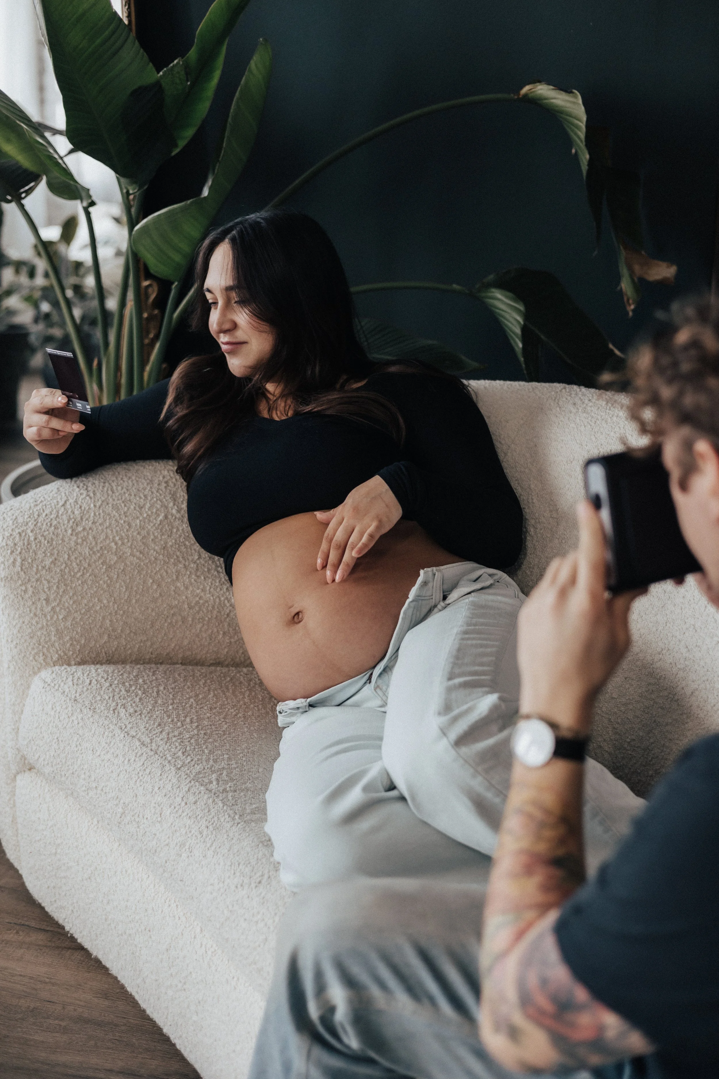 Woman relaxing on a cream-colored sofa, lifting her shirt to reveal her pregnant belly, while a man with tattoos takes a photo of her with an instant camera in Cleveland Ohio