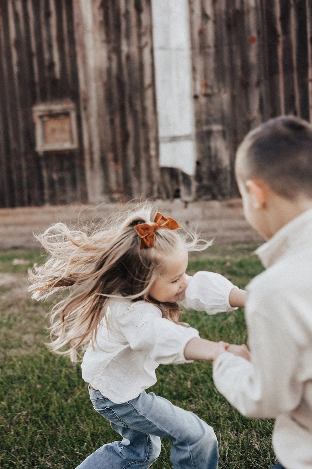 Two children, a girl with blonde hair and a boy with dark hair, playing and holding hands on a grassy yard with a wooden barn in the background.