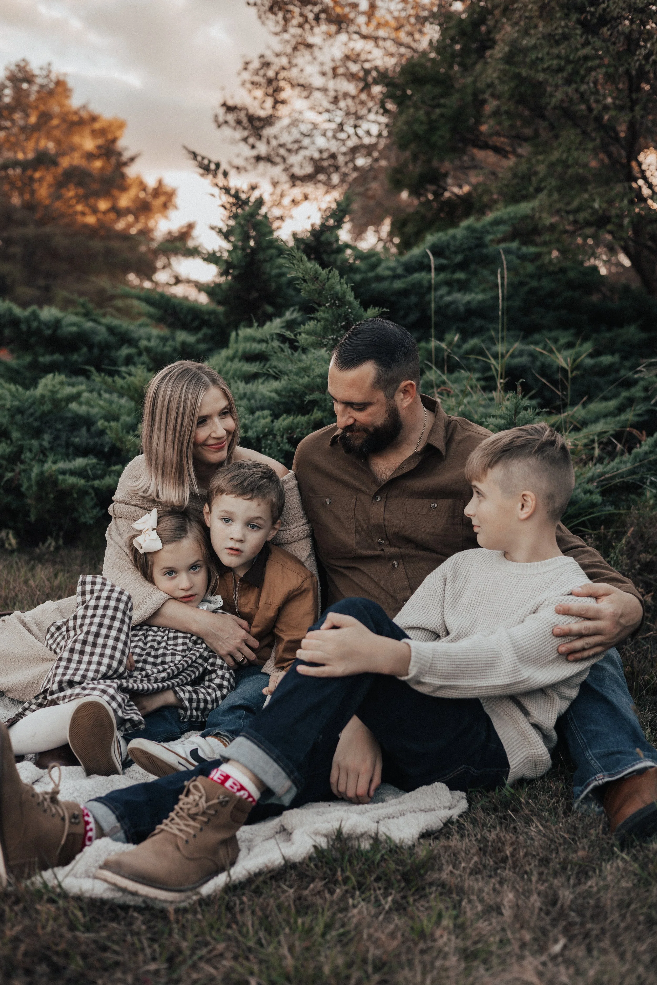 A family of five sitting outdoors on a blanket among evergreen trees during sunset. The mother and father are cuddling with their three children, smiling and enjoying the moment.